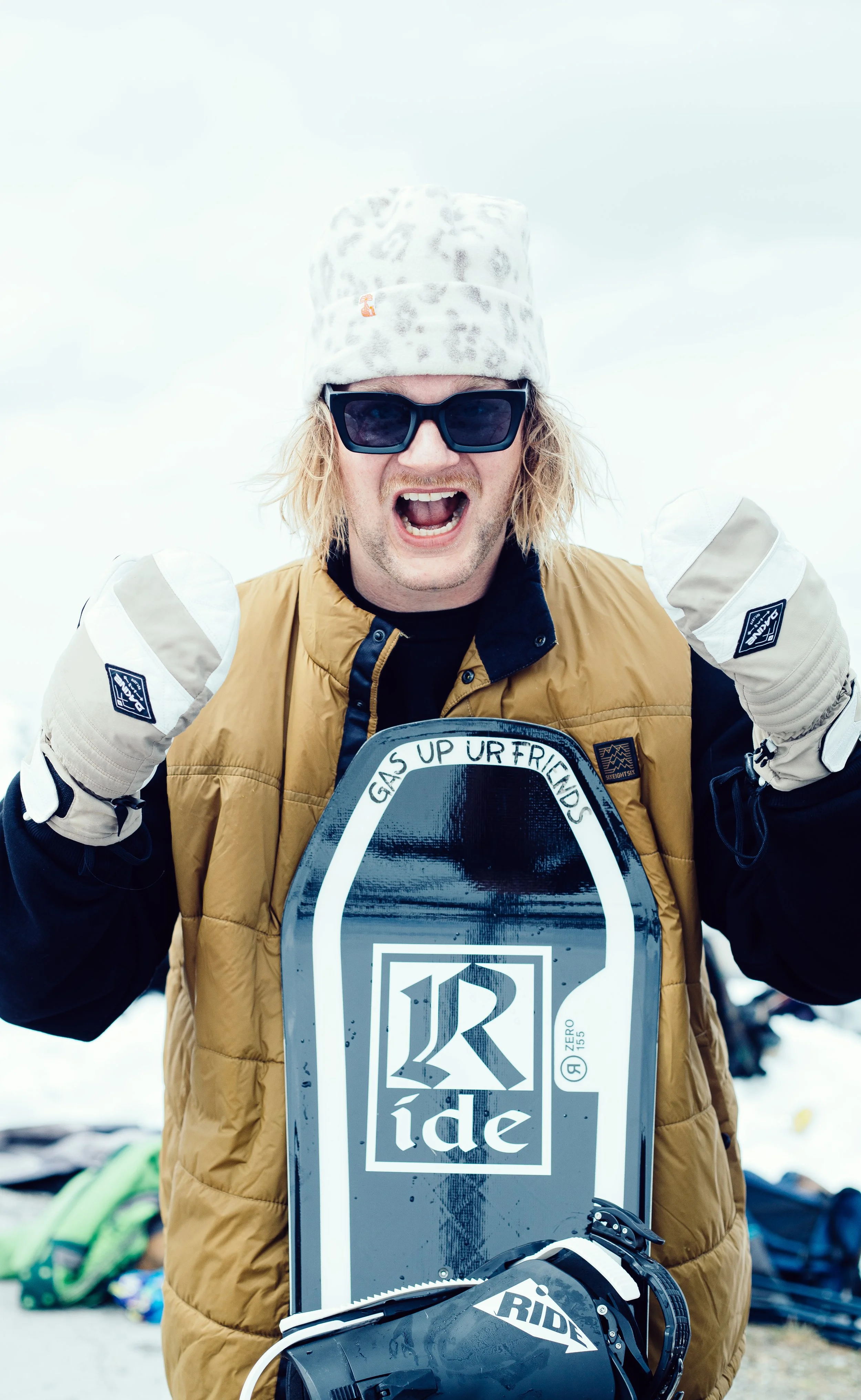 Ein Mann trägt einen Leopardenmuster-Pullover, eine beige Weste, schwarze Sonnenbrille und Handschuhe, steht im Schnee, jubelt und posiert mit einem Snowboard.
