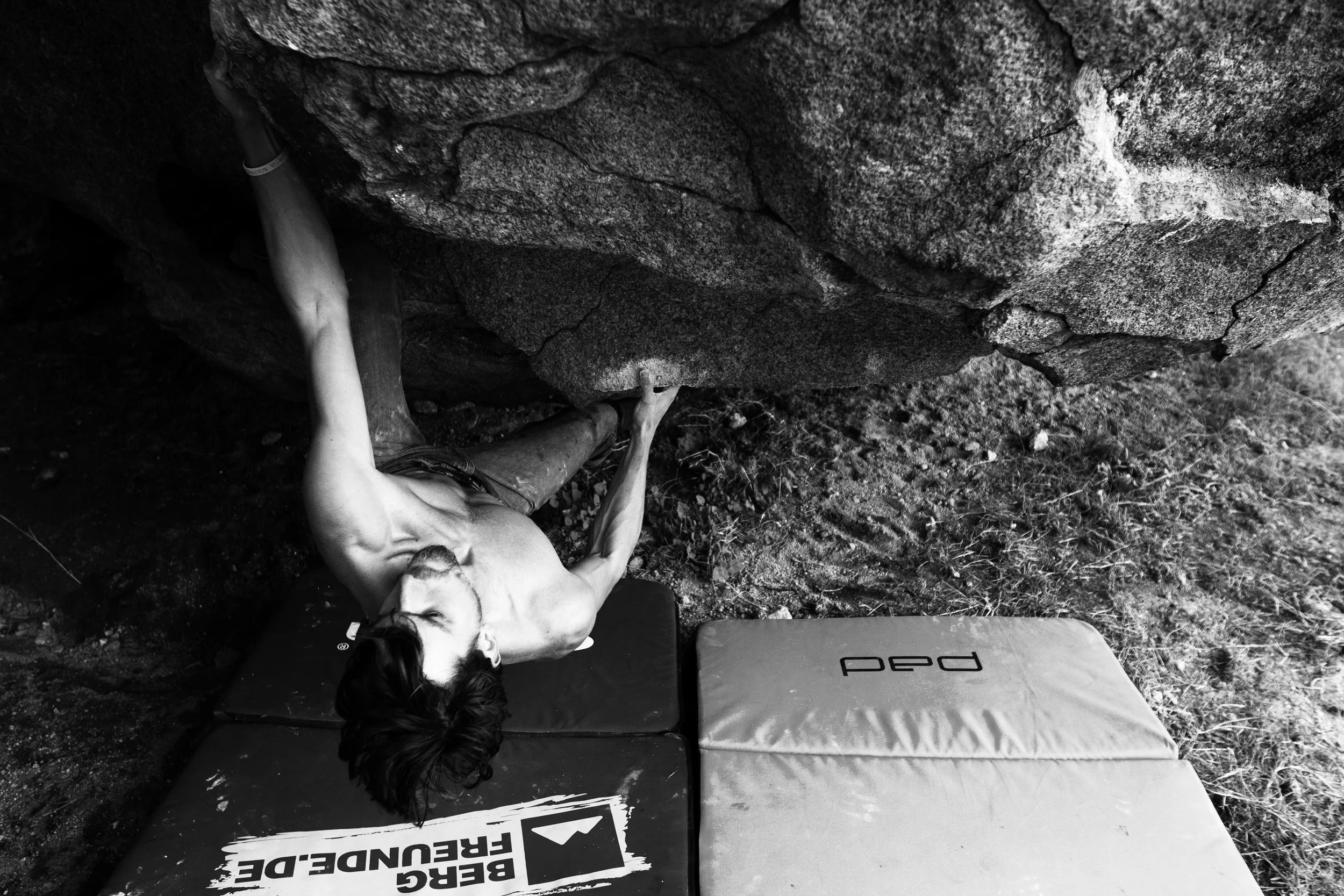 Mann klettert auf einer Felswand, liegt auf einer Matratze, trägt keine Schuhe, mit dunklen Haaren und Bart, in einer natürlichen, felsigen Umgebung, schwarz-weiß