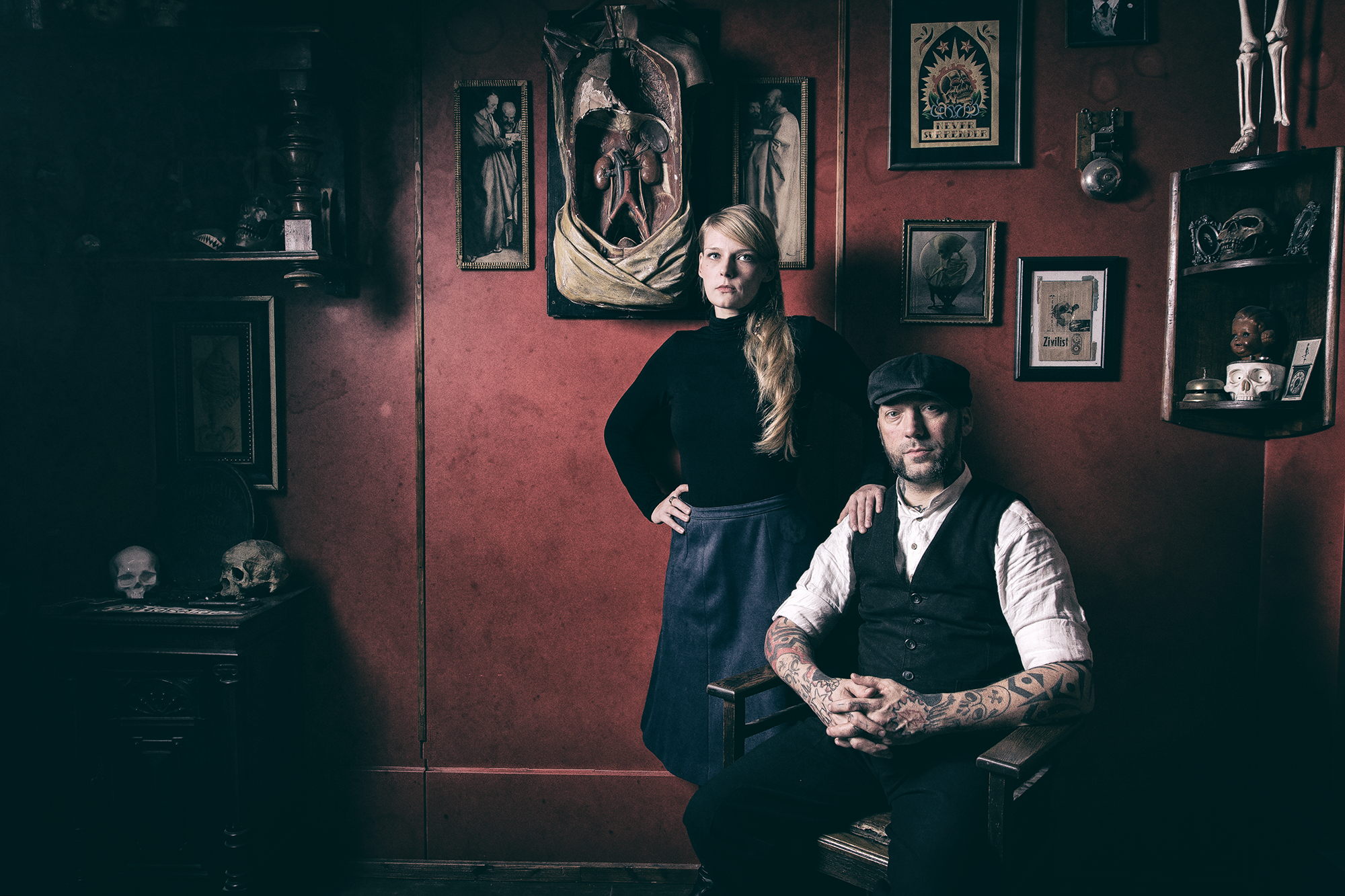 Ein Mann sitzt auf einem Stuhl mit Tattoos auf den Armen, trägt eine schwarze Weste und einen schwarzen Hut. Eine Frau steht dahinter mit einer Hand auf seiner Schulter, trägt ein schwarzes Oberteil und eine lange dunkelblaue Rock. Der Raum ist mit d