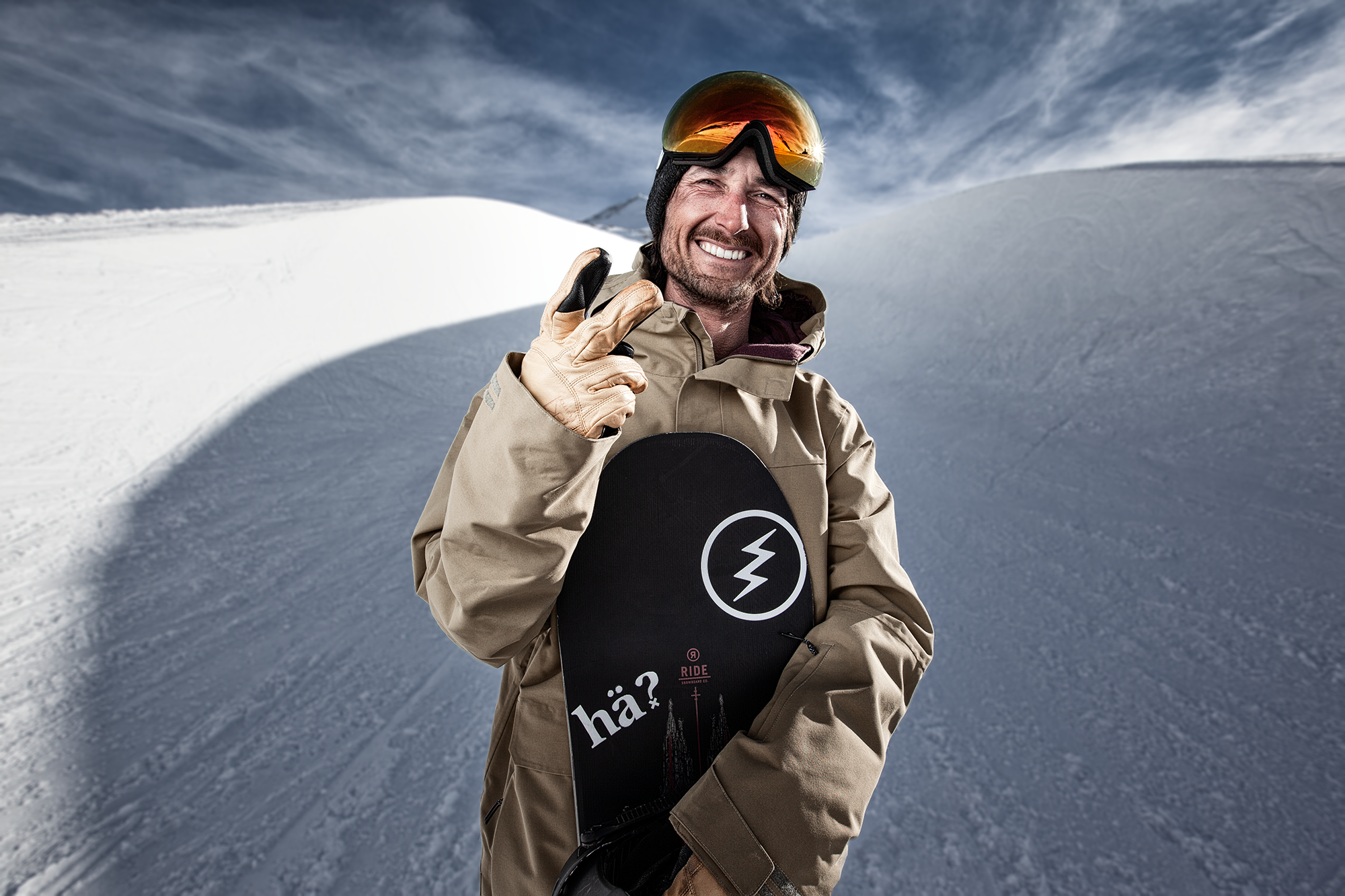Ein lachender Skifahrer trägt eine braune Jacke, einen Helm mit Schutzbrille, Gleitpads, die einen Blitz symbolisieren, und hält ein Snowboard mit der Aufschrift 'hää?'. Er steht vor einer schneebedeckten Berglandschaft unter einem bewölkten Himmel.