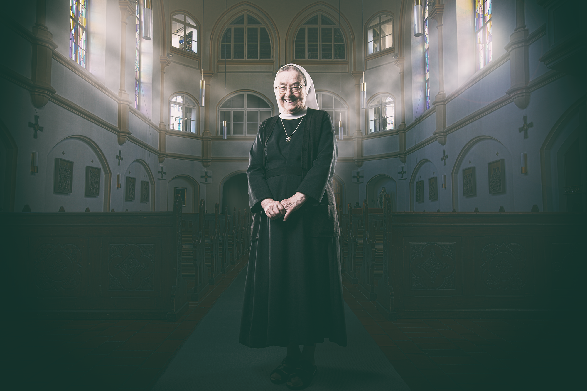 Portrait einer lächelnden Nonne in einer Kirche mit hohen Fenstern und Sonnenlicht, das durch bunte Glasfenster einfällt. Chris Reichl Portraits.