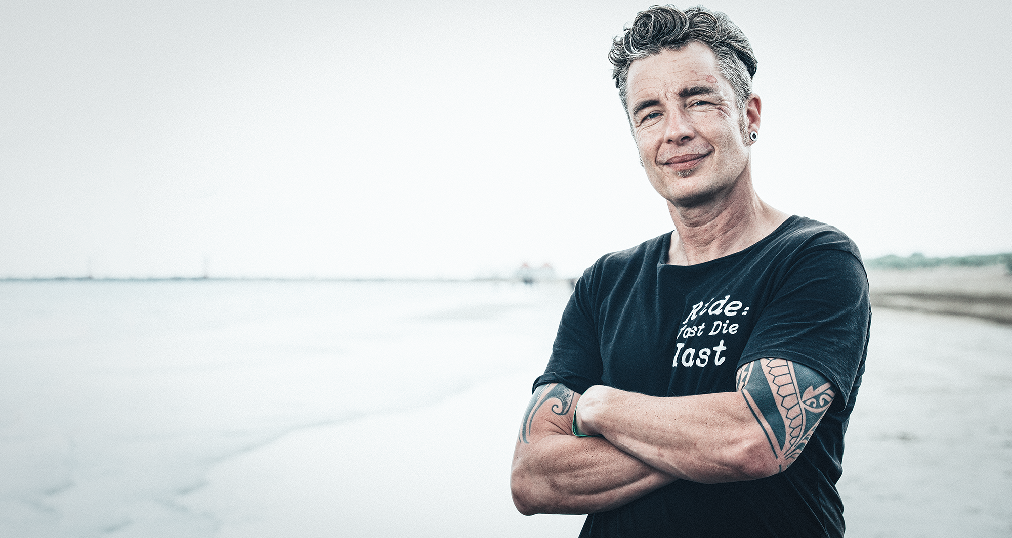 Ein Mann steht am Strand mit verschränkten Armen und lächelt. Er trägt ein schwarzes T-Shirt mit weißem Text und hat Tattoos am Arm.