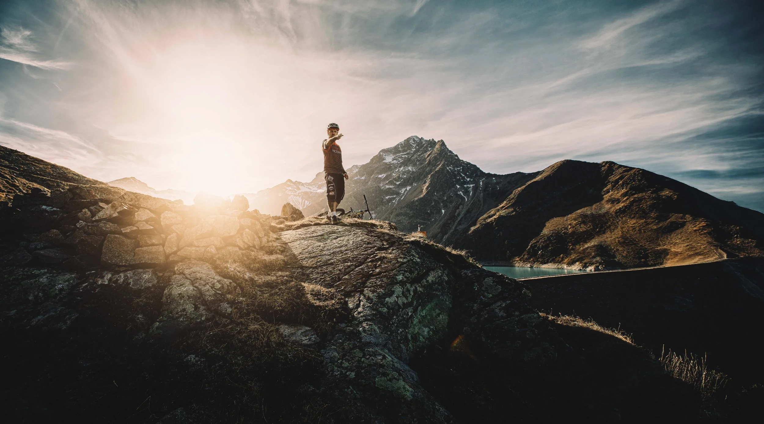 Ein Mann steht auf einem Felsen in den Bergen bei Sonnenuntergang, mit einem Fahrrad am Boden neben ihm, Blick auf einen See und schneebedeckte Gipfel im Hintergrund.
