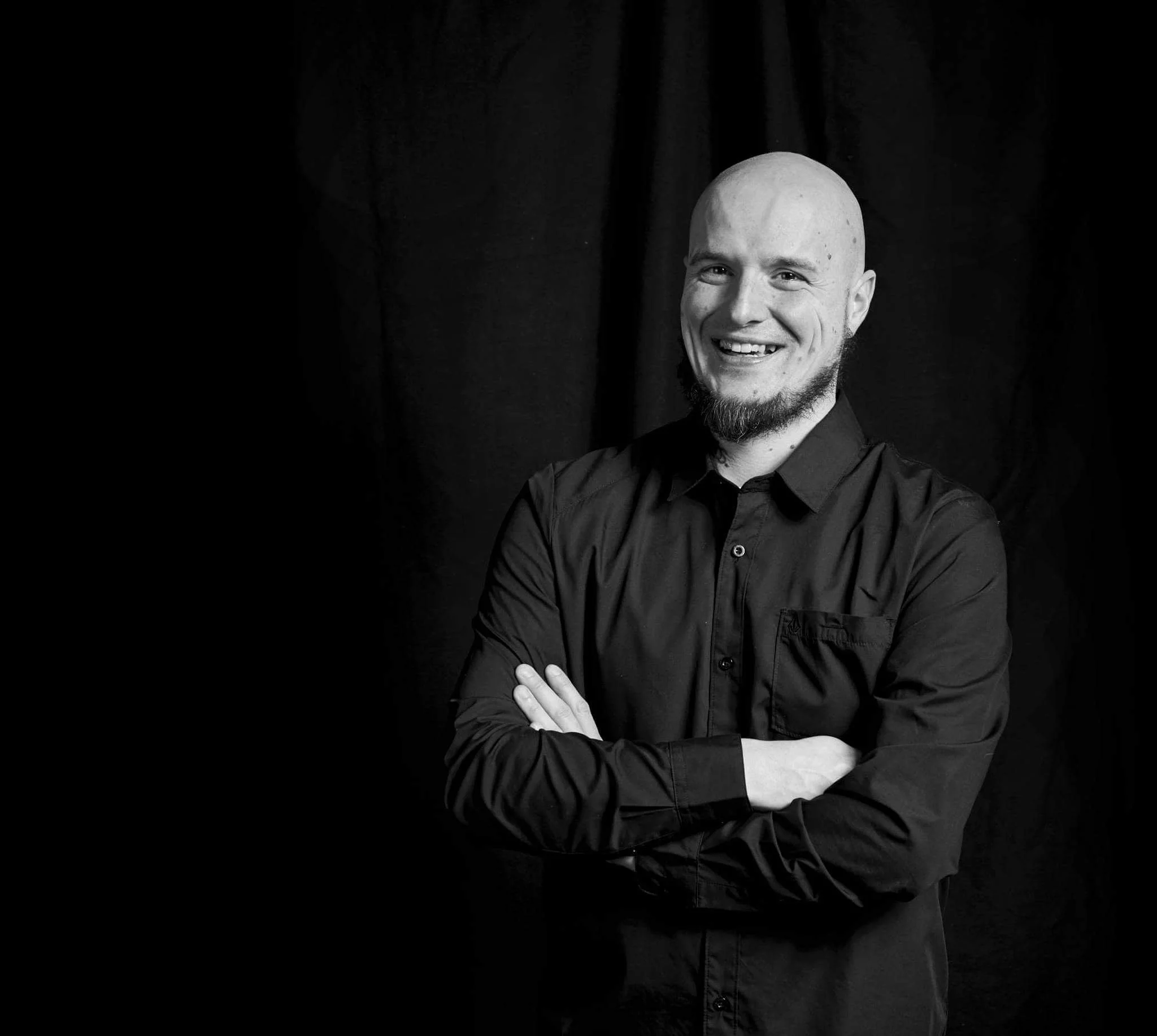 Schwarz-weiß Portrait eines lachenden, bärtigen Mannes mit Glatze, der schwarze Kleidung trägt und die Arme verschränkt hat, vor dunklem Hintergrund. Selbstportrait Chris Reichl.