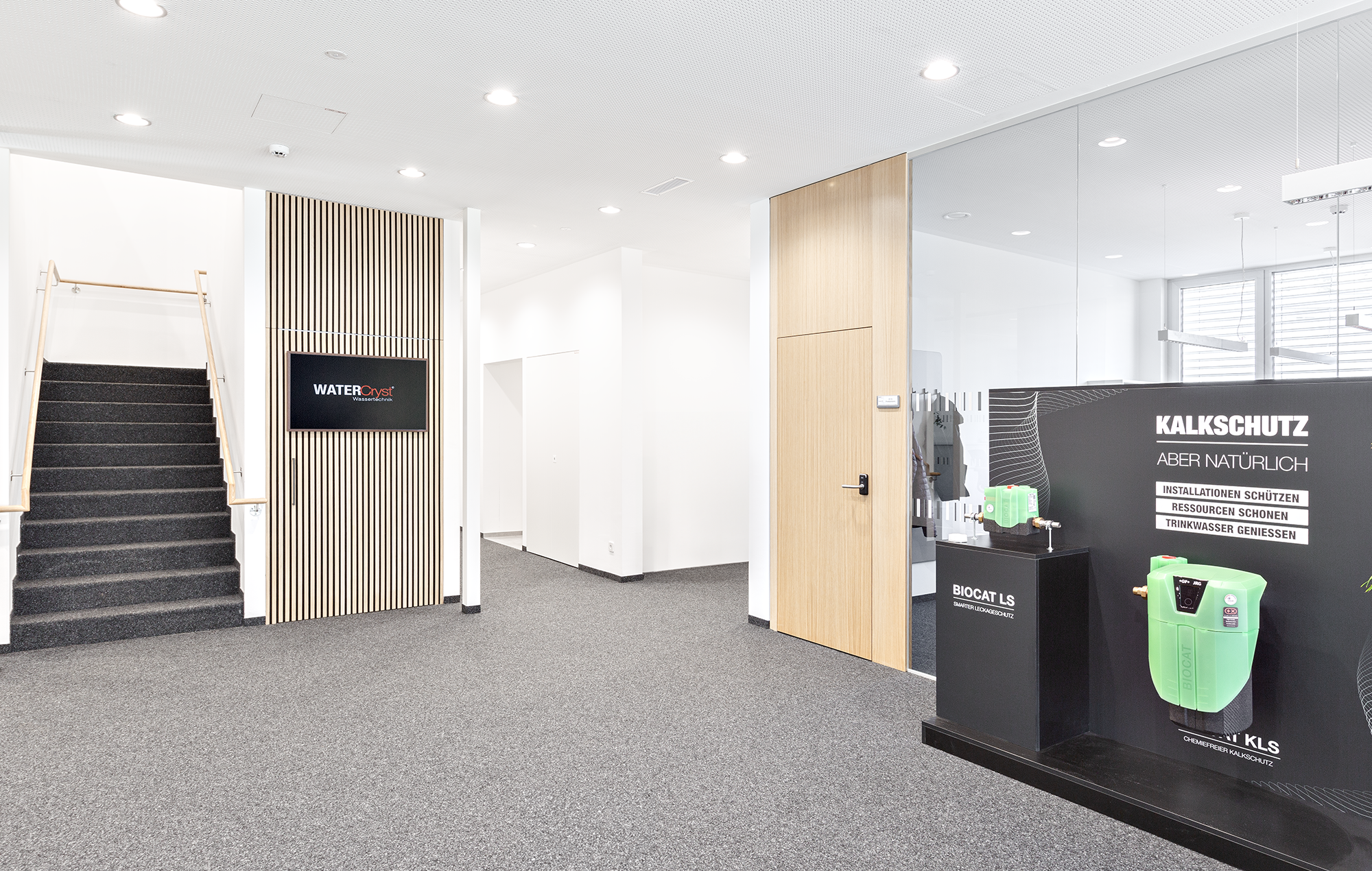 Innenaufnahme eines modernen Büros mit Treppe, Wandbildschirm und einer Ausstellung zu Kalkschutztechnik, inklusive Geräte und Infotafeln. Chris Reichl Architektur.