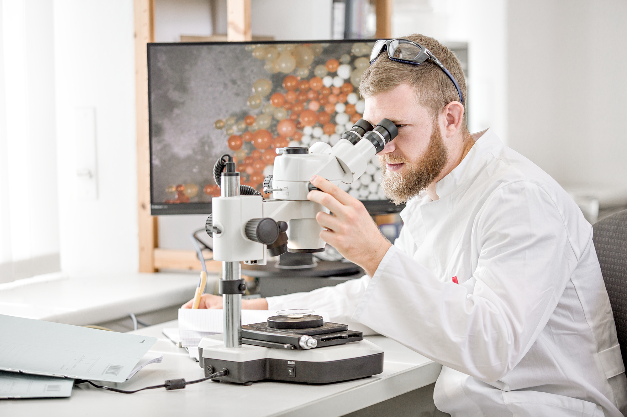 Ein Mitarbeiter blickt durch ein Mikroskop in einem Labor, im Hintergrund ein Bildschirm mit einer Moleküldarstellung. Chris Reichl on Location Shots.