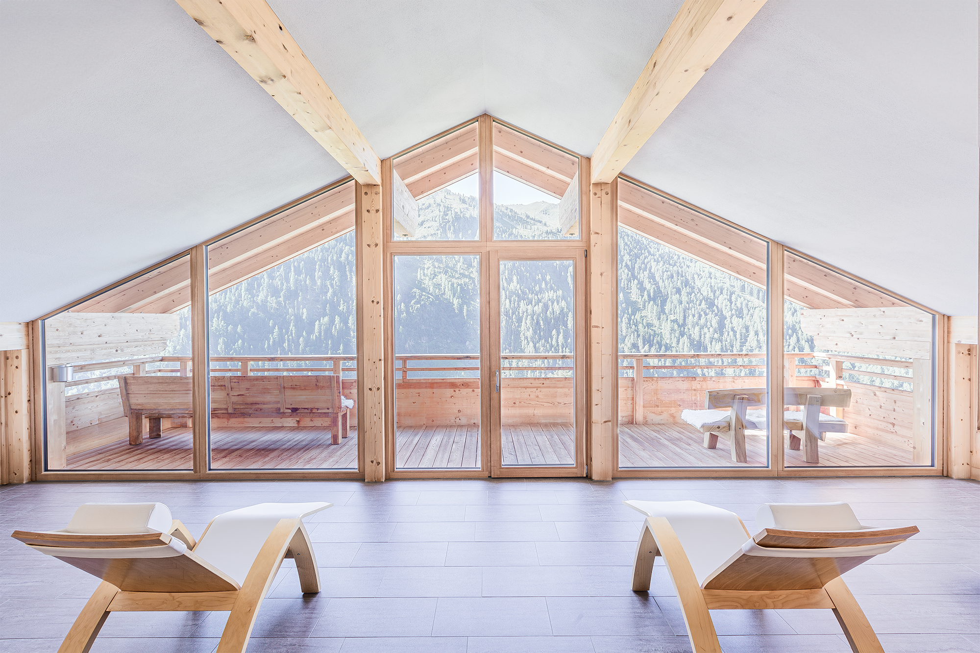 Innenraum mit Holzrahmen und großen Fenstern, Blick auf Berge, zwei Holz-Liegestühle, Holz-Sitzbank und Holz-Tisch auf Balkon. Chris Reichl Architektur Shots.