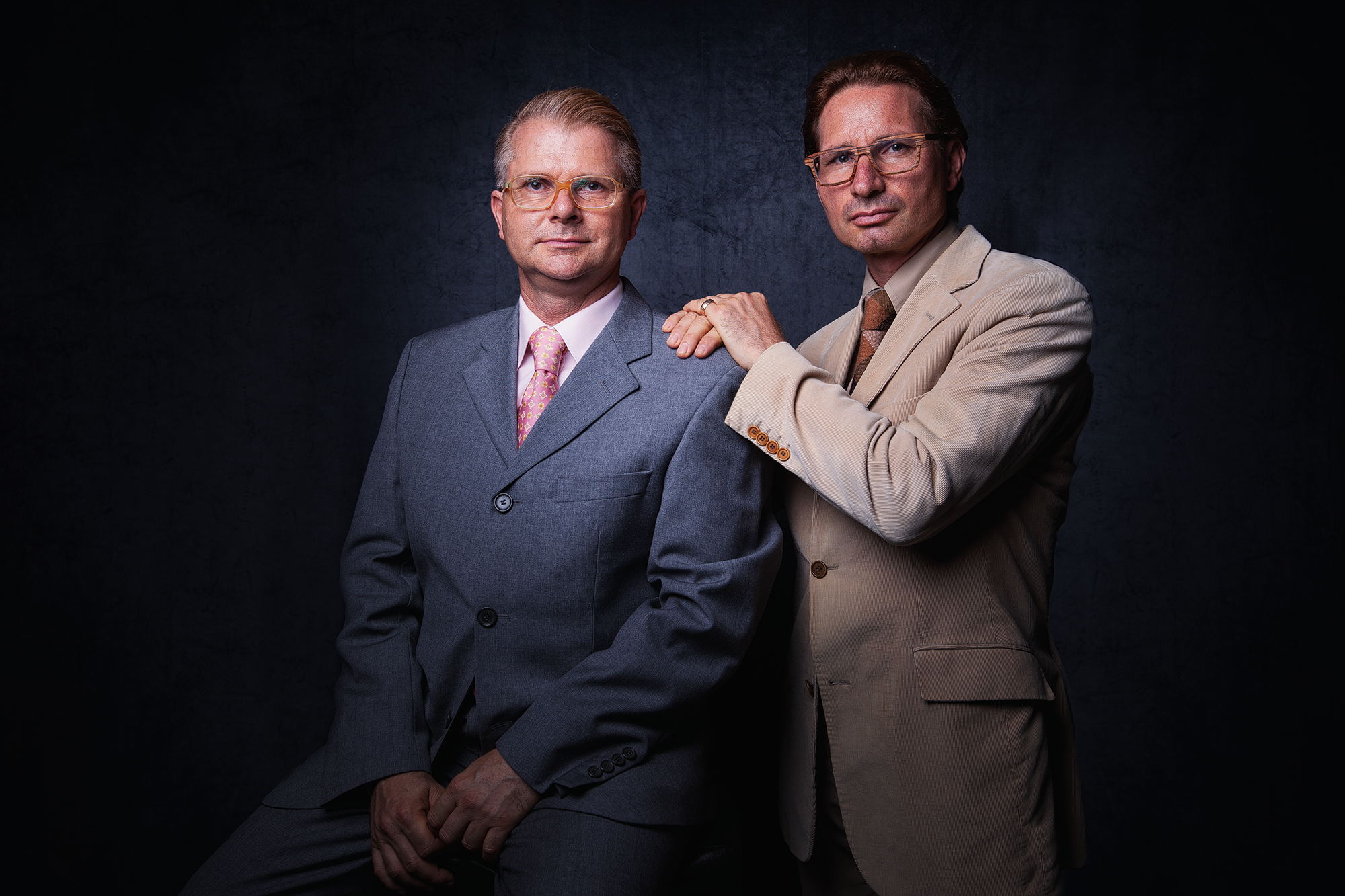 Zwei gut gekleidete Männer in Anzügen posieren vor dunklem Hintergrund, einer sitzt, der andere steht dahinter mit Hand auf Schulter des Sitzen. Businessportraits von Chris Reichl.