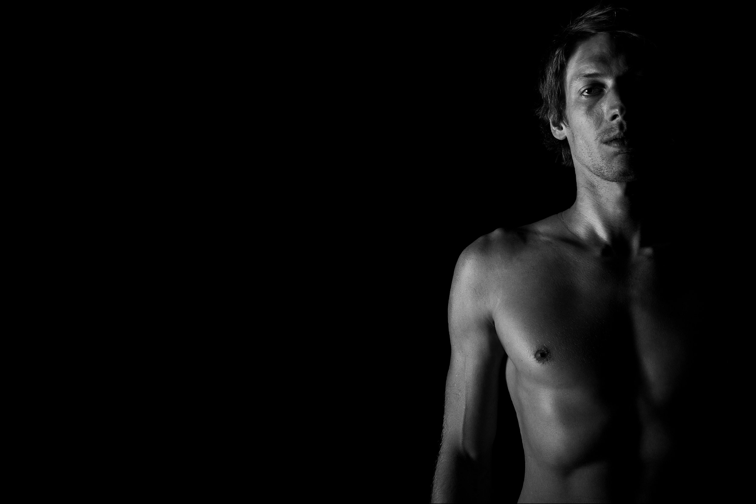 Schwarz-weiß-Foto eines nackten jungen Mannes, der nur zur Hälfte im Bild zu sehen ist, mit dunklem Hintergrund.