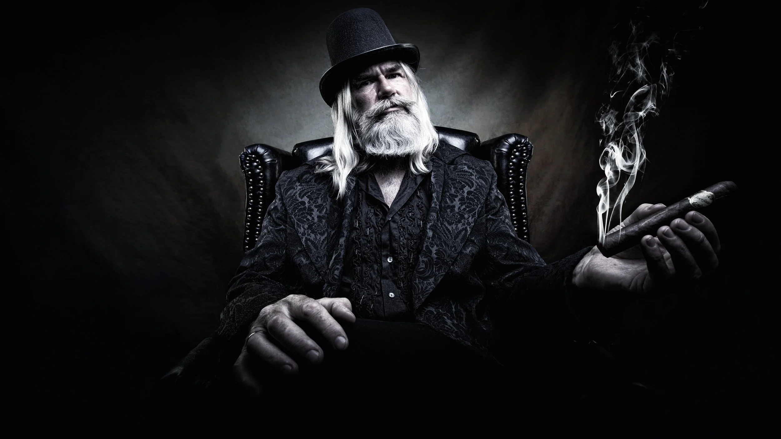 Ein alter Mann mit langem weißem Bart und Haaren sitzt in einem schwarzen Sessel und raucht eine Zigarre, umgeben von Dunkelheit.