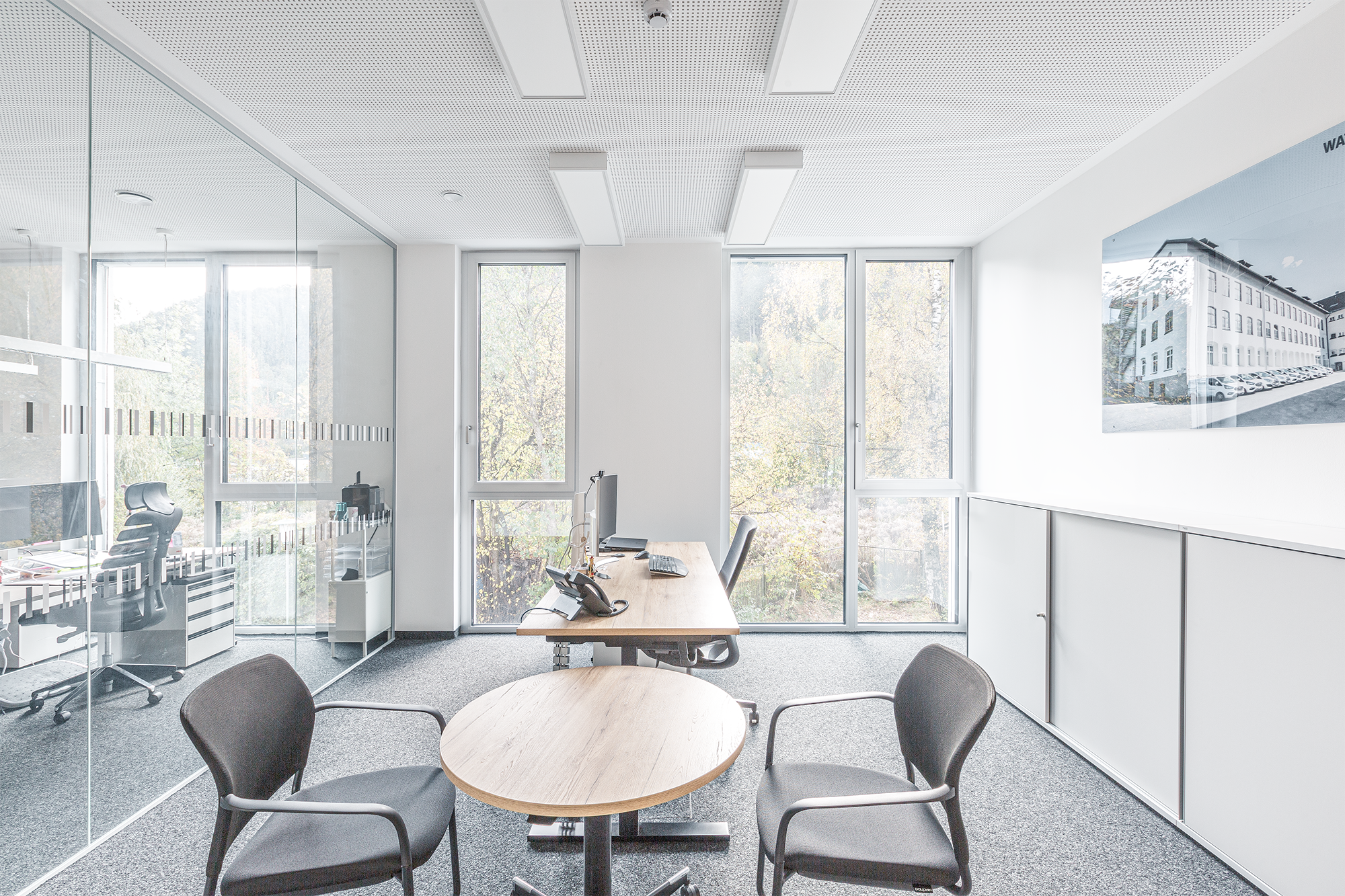 Leeres Büro mit Schreibtisch, Computer, Telefon, zwei Stühlen und einem großen Fenster mit Blick nach draußen.