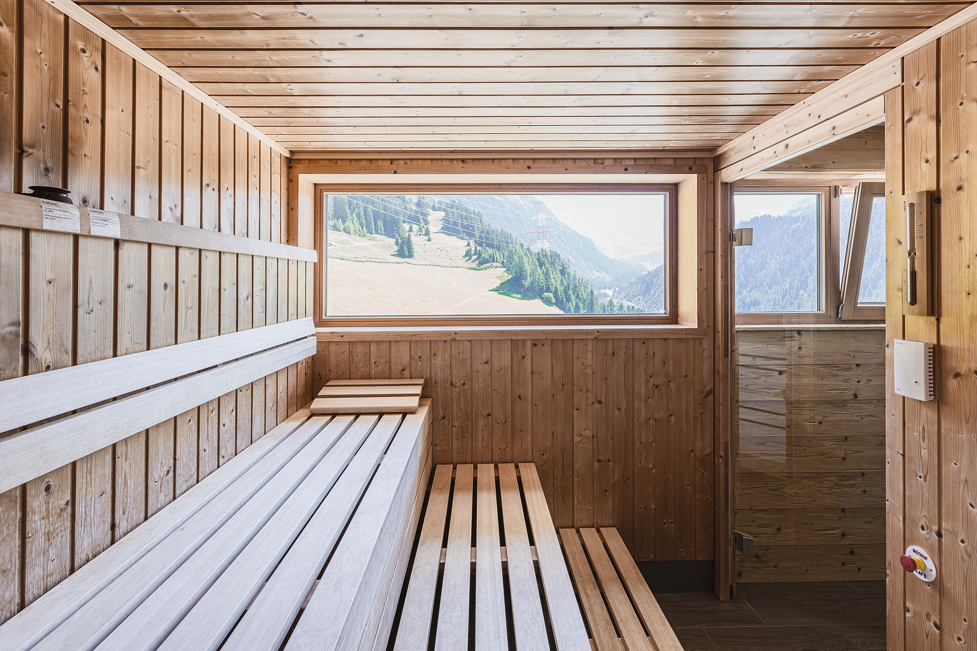 Innenraum einer Holzsauna mit großen Fenstern, die eine Berglandschaft zeigen, Holzbänken und Holzverkleidung.