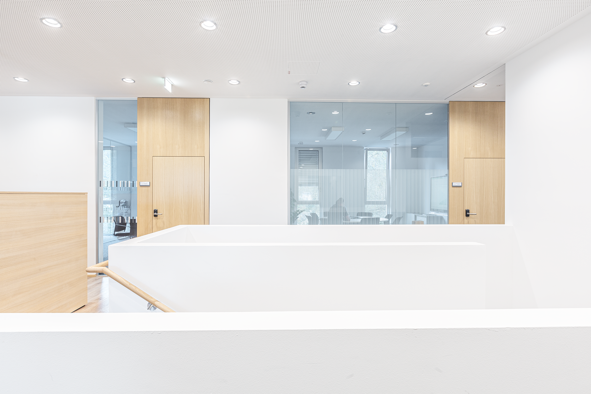 Innenansicht eines modernen, hellen Büros mit weißen Wänden, Holzschiebetüren und einer gläsernen Besprechungsraumwand. Es gibt Deckenleuchten, und im Hintergrund sind Fenster sichtbar.