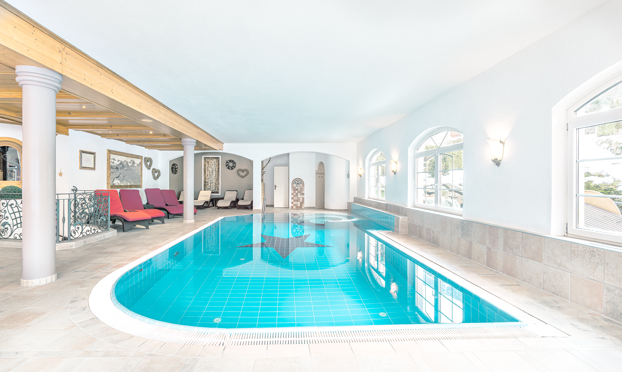 Innenansicht eines warmen Hallenbads mit blauer Fliesenbadewanne, Liegen und großen Fenstern, weiße Wände und Dekorationsherzen auf der Wand.