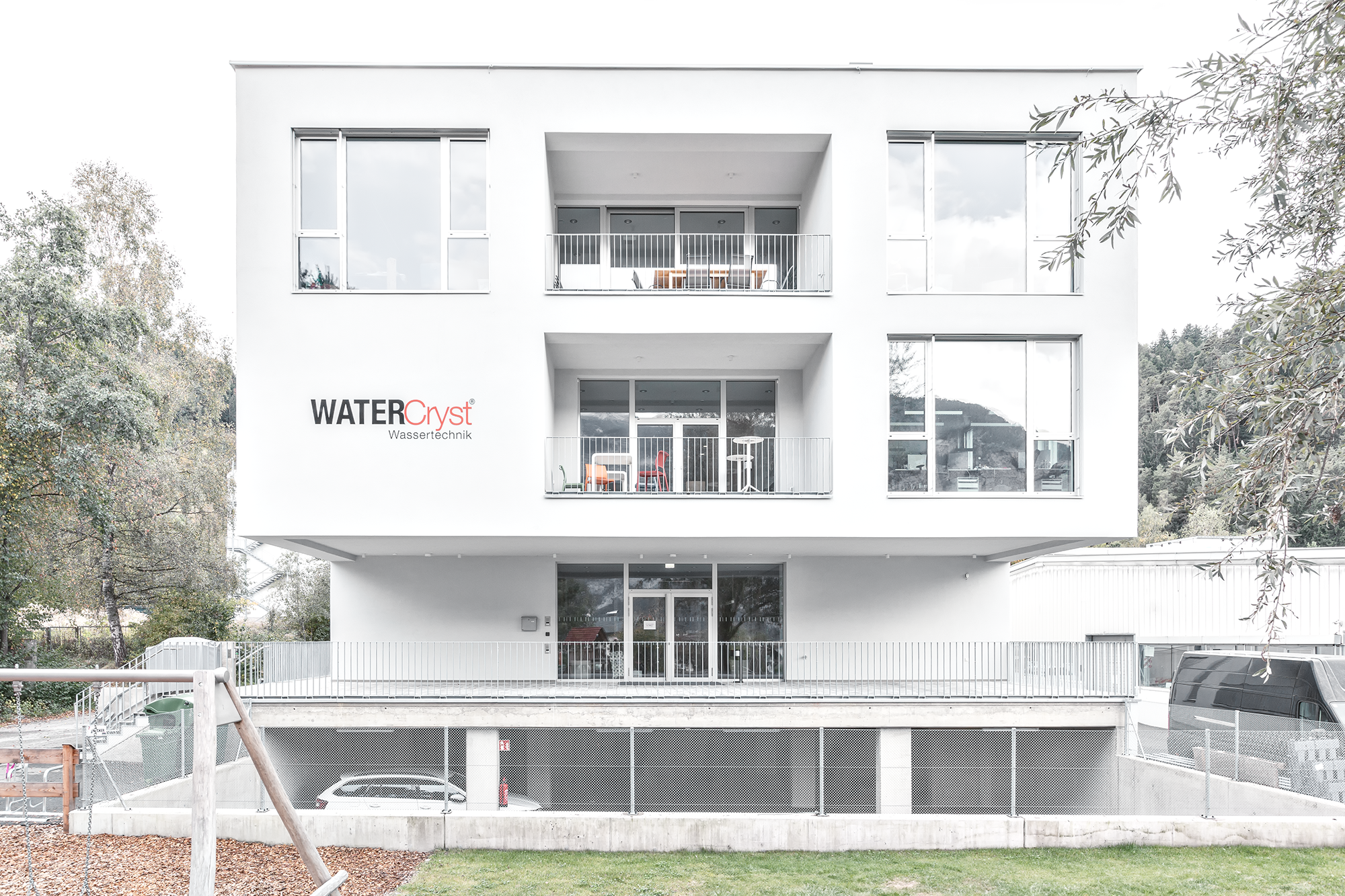 Mehrstöckiges weißes Gebäude mit Balkon und Fenstern, außen mit Firma 'WATER Cryst Wassertechnik' beschriftet, im Hintergrund Bäume und Hügel, im Vordergrund Rasen und Spielgerät.
