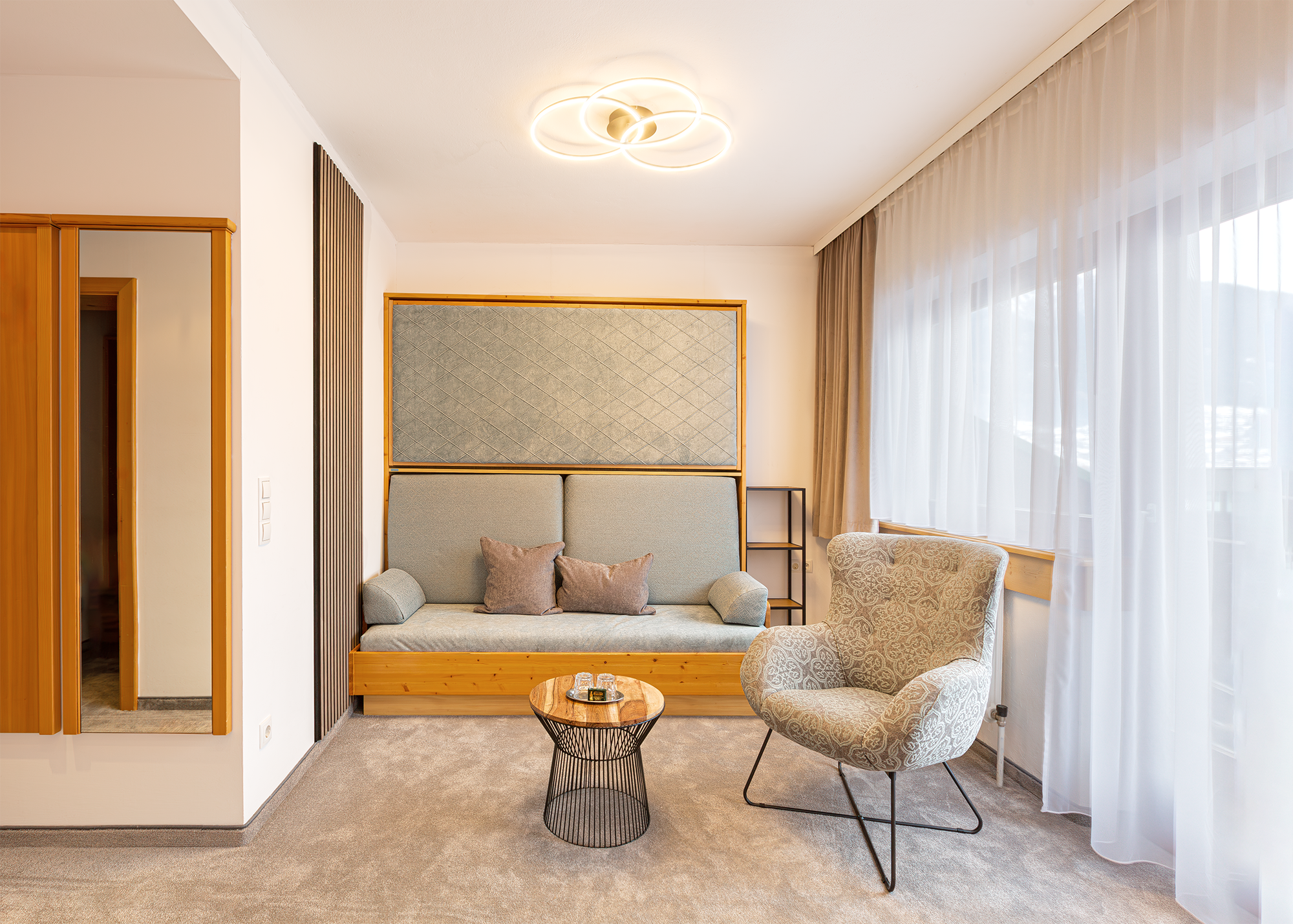 Ein modern eingerichteter Wohnbereich mit einem grauen Sofa, einem gepolsterten Sessel und einem kleinen runden Beistelltisch vor einem großen Fenster mit weißen Vorhängen.