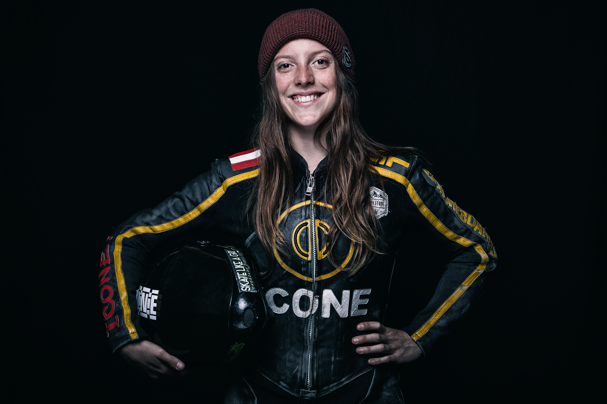 Junge Frau in Motorsport-Overall mit Helm in der Hand, lächelnd, dunkler Hintergrund.