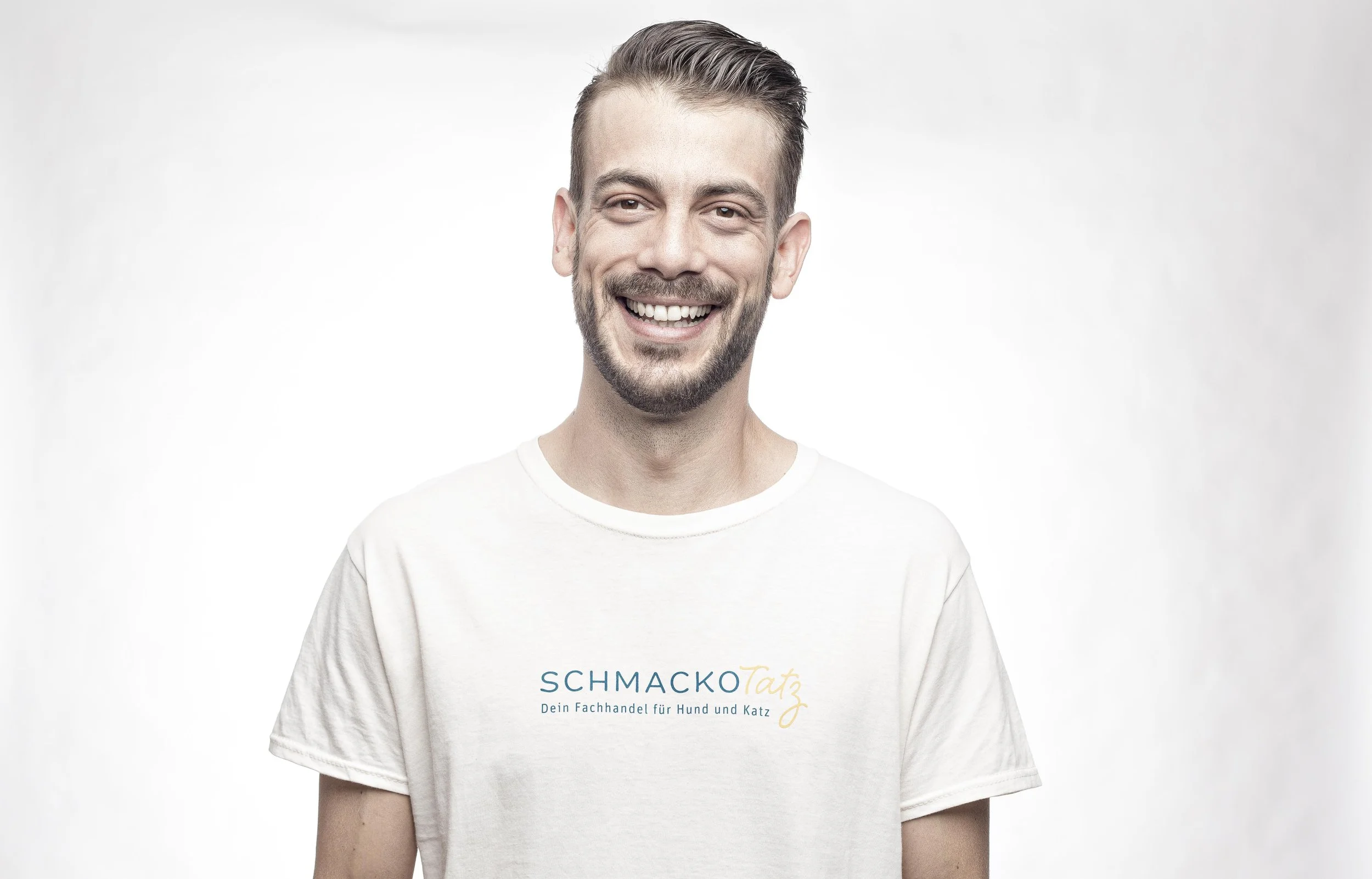 Junger Mann mit Bart und Kurzhaarschnitt, lächelnd, trägt weißes T-Shirt mit Logo "SchmackoTatz" auf weißem Hintergrund.