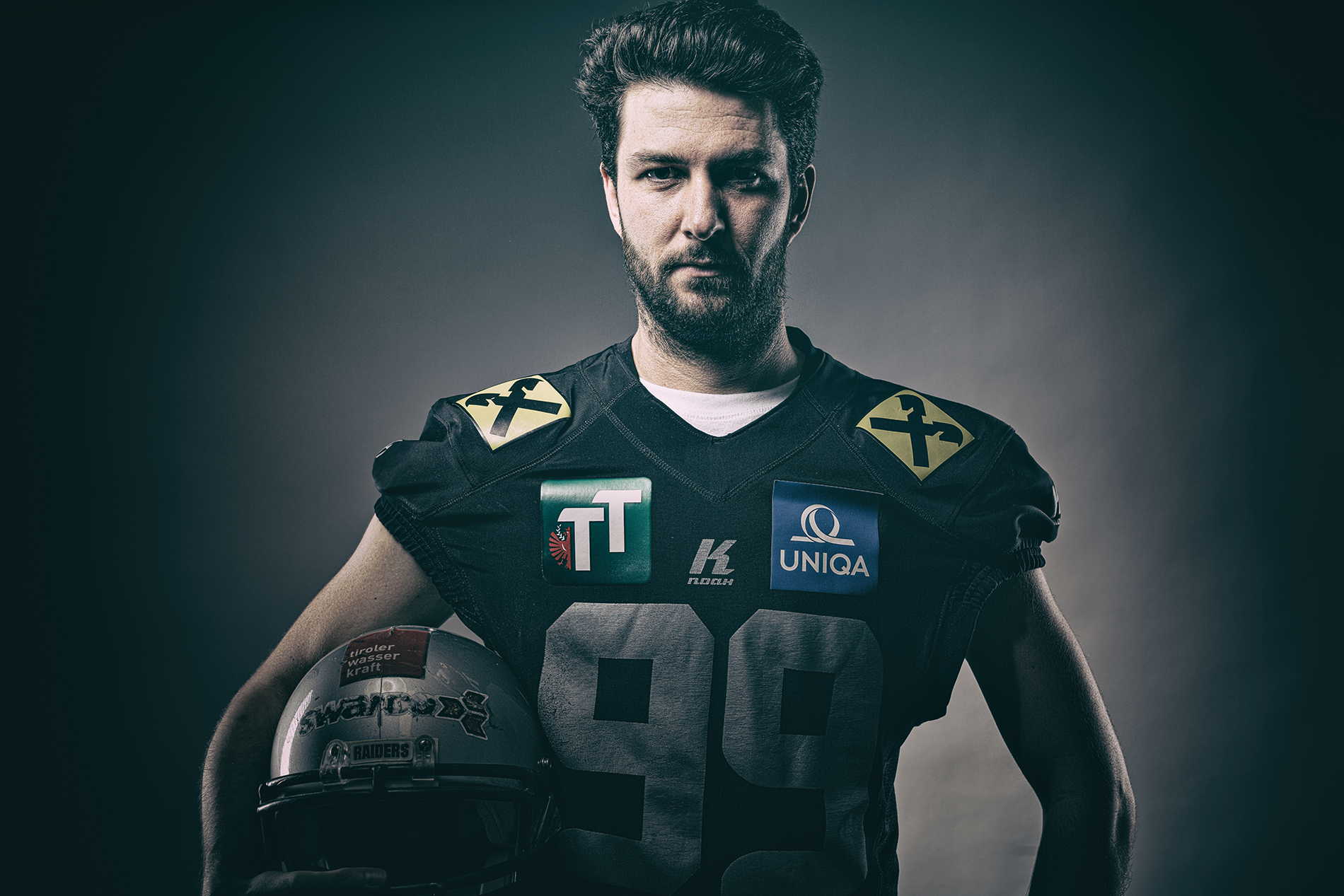 Ein männlicher American Football-Spieler in schwarzer Uniform hält einen Helm in der Hand, steht vor dunkelgrauem Hintergrund und blickt ernst in die Kamera.
