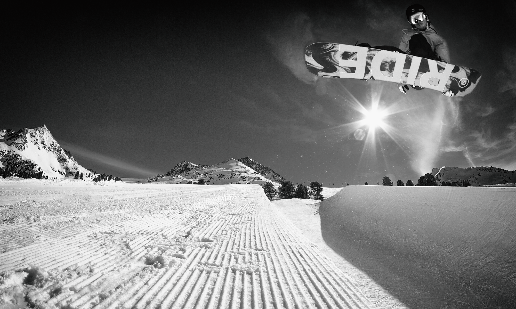 Ein Snowboarder springt in der Luft auf einem verschneiten Skigelände bei Sonnenlicht, im Hintergrund Berge und Schnee. Schwarz-Weiß-Bild.
