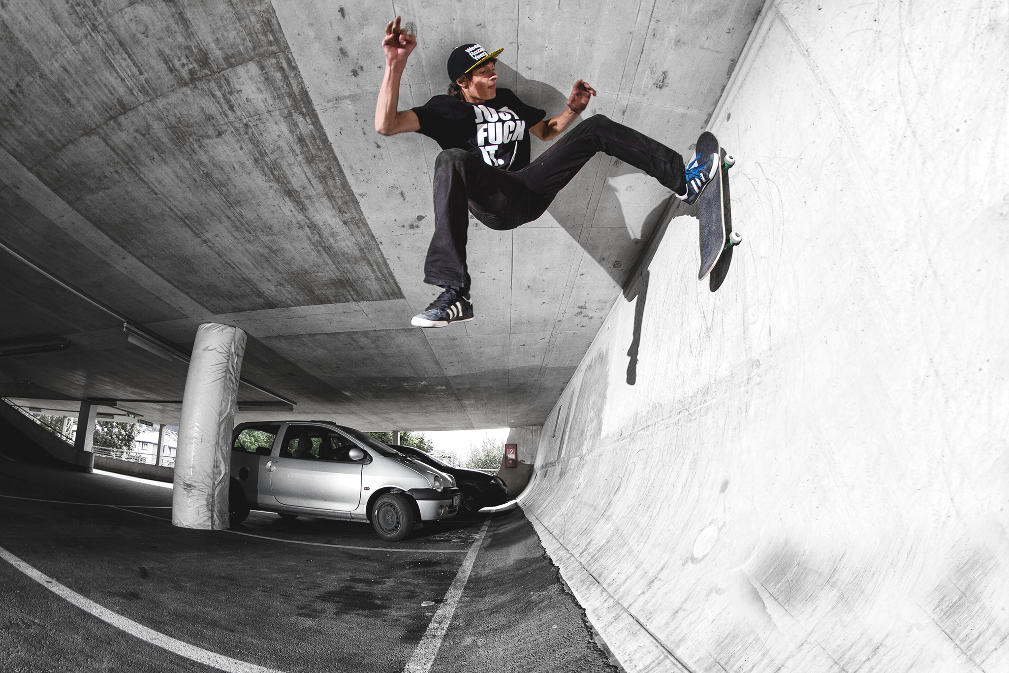 Junger Skateboarder in einem Parkhaus, der eine Wand entlang fährt, trägt ein schwarzes T-Shirt mit weißen Texten und eine schwarze Cap.