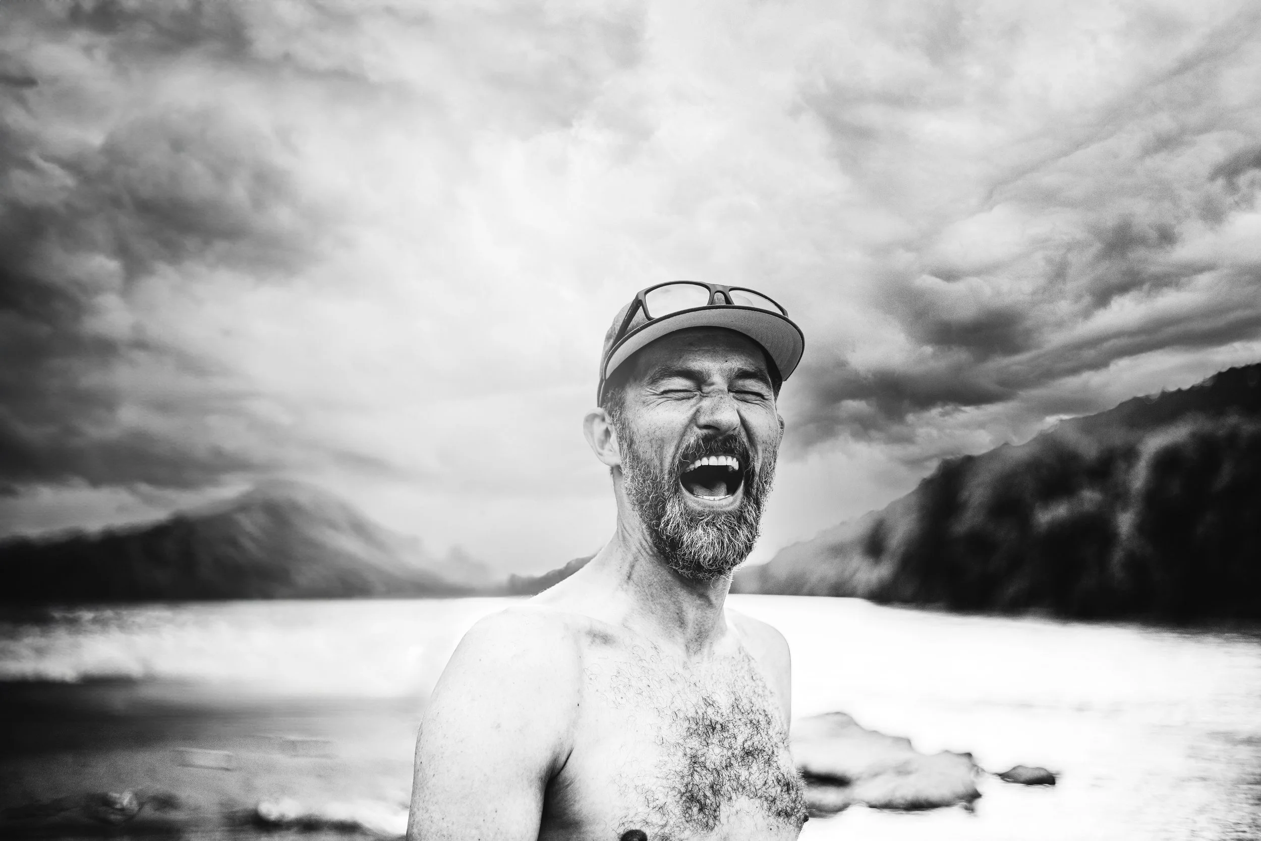 Schwarz-weiß Foto eines lachenden Mannes mit Bart, der keine Oberbekleidung trägt, am Strand mit Bergen und Wolken im Hintergrund.