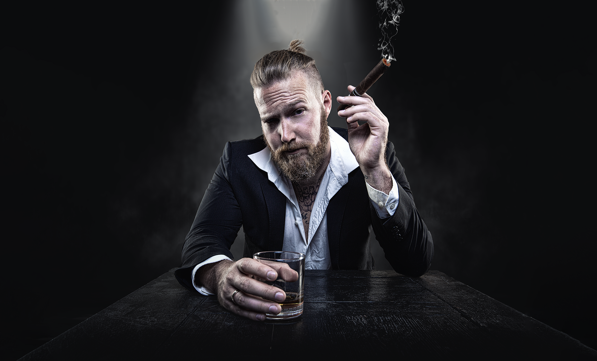 Ein Mann mit Bart, angezogen in einem schwarzen Anzug und weißem Hemd, sitzt an einem dunklen Tisch, hält ein Glass Whiskey in der Hand und raucht eine Zigarre, vor dunklem Hintergrund.