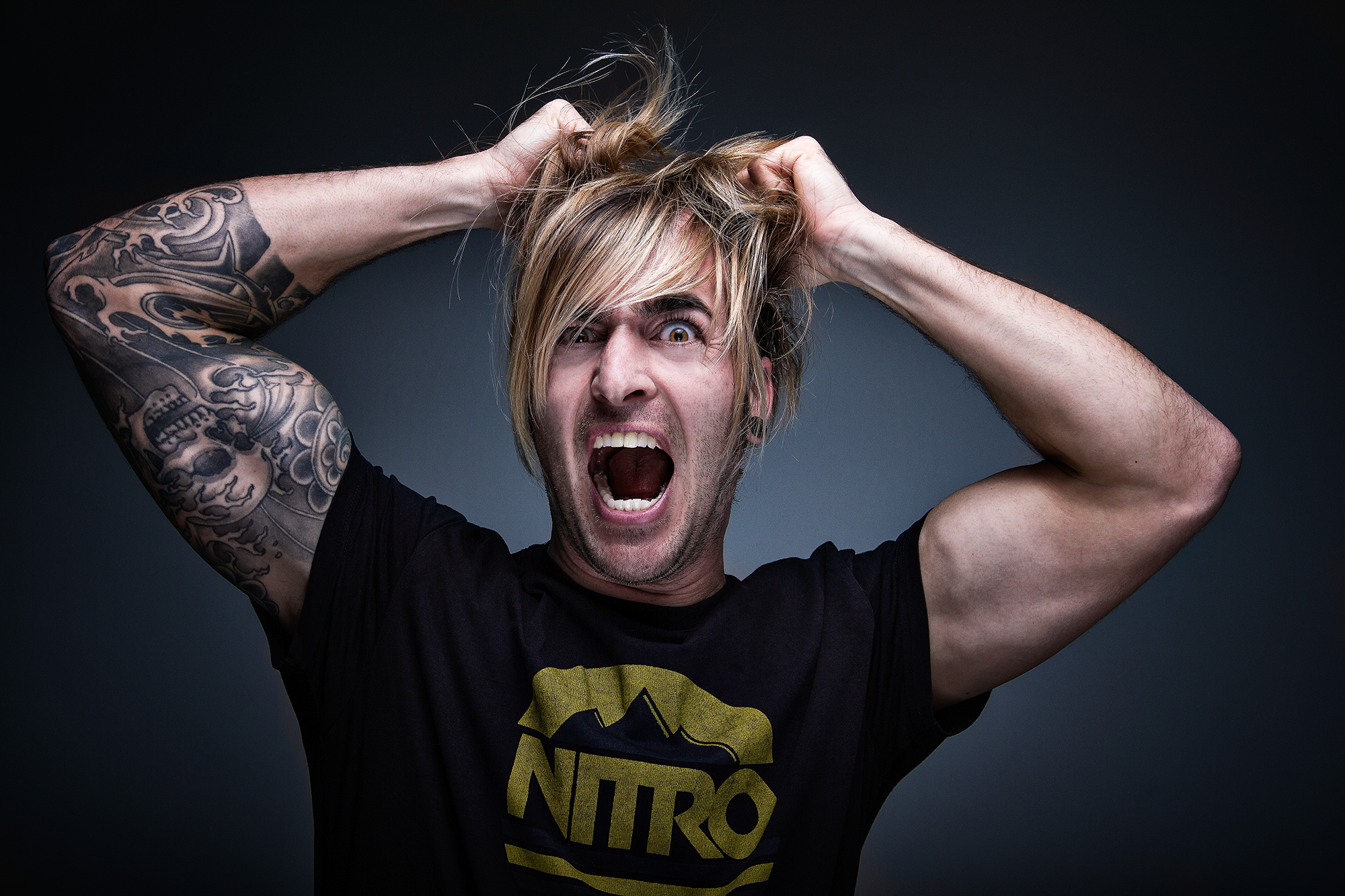 Ein emotionale Mann schreit mit offenem Mund, trägt ein schwarzes T-Shirt mit einem gelben Logo, hat lange blonde Haare, zeigt Tattoos am linken Arm, hält seine Haare mit beiden Händen, vor dunklem Hintergrund.