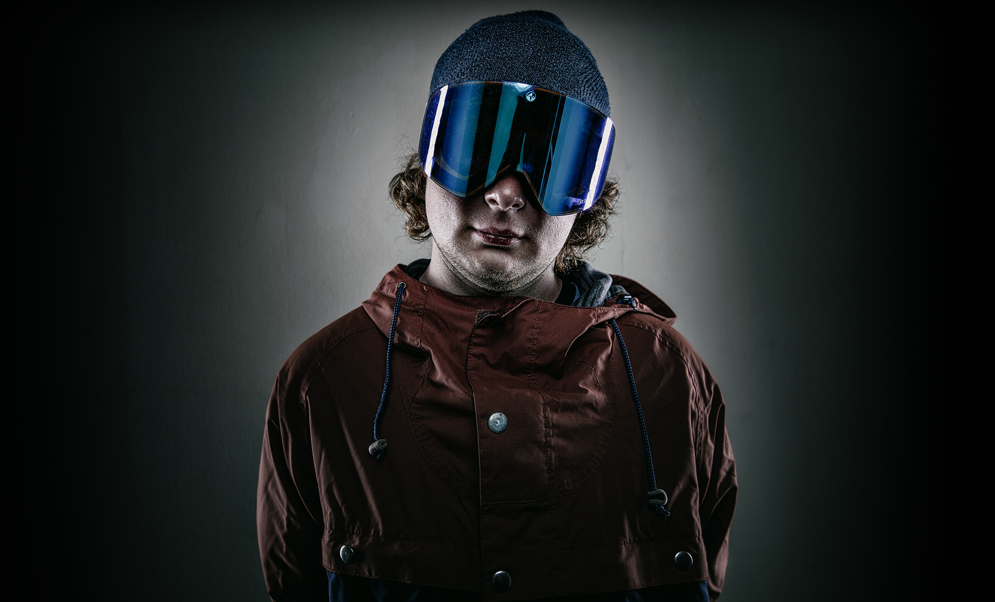 Mann mit Kapuzenjacke und Skibrille vor dunklem Hintergrund.