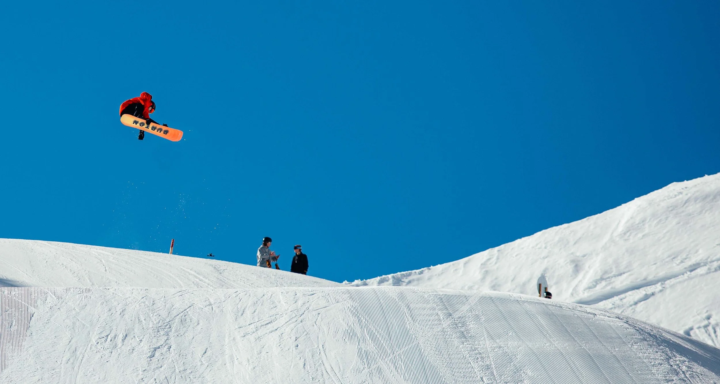 Skifahrer springt mit Snowboard in die Luft auf einer verschneiten Piste bei klarem Himmel, während zwei Personen im Hintergrund zuschauen.
