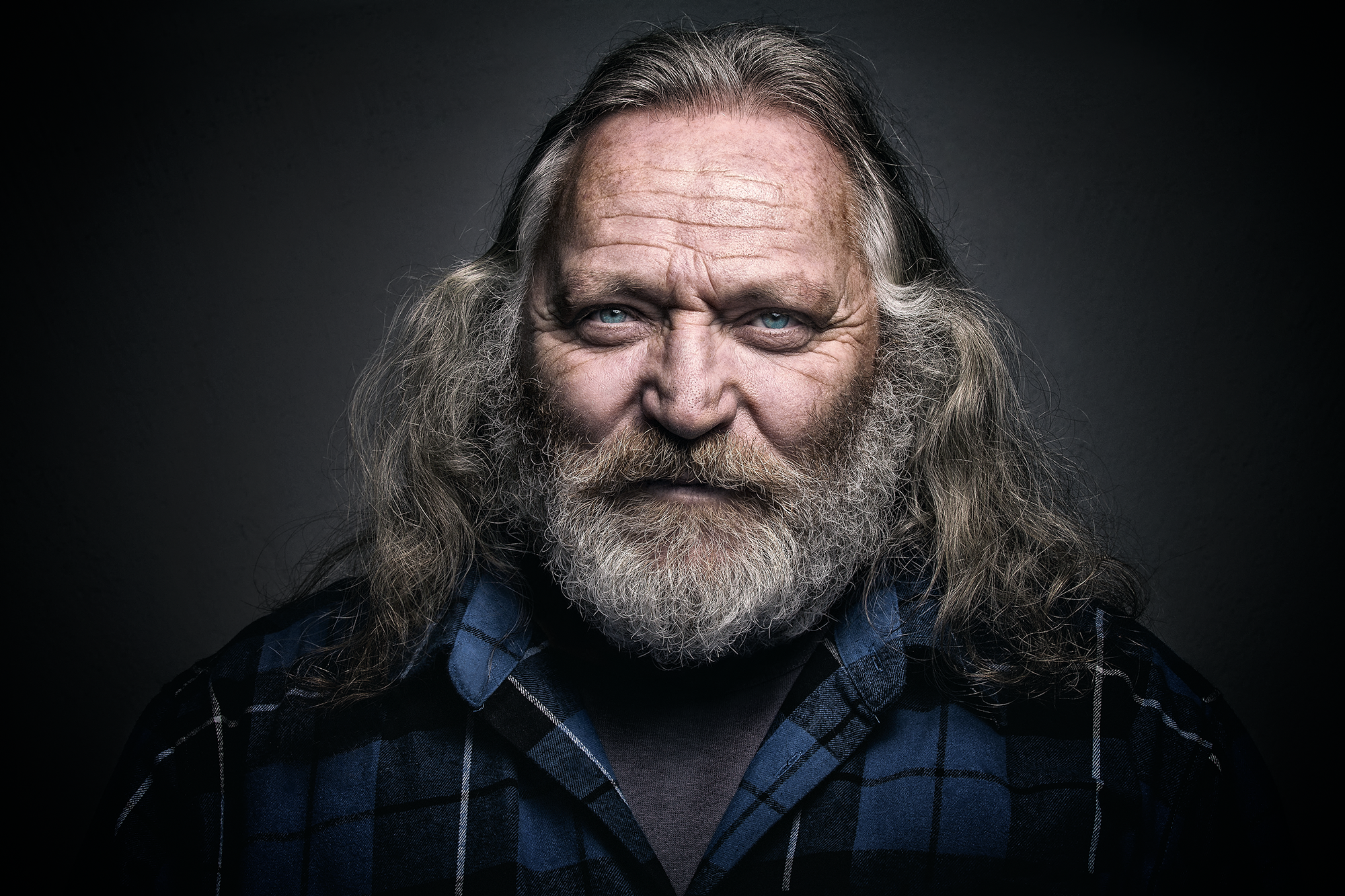 Porträt eines älteren Mannes mit grauem, langen, gewellten Haar und Bart, trägt eine dunkle, blau-karierte Hemd, dunkler Hintergrund.