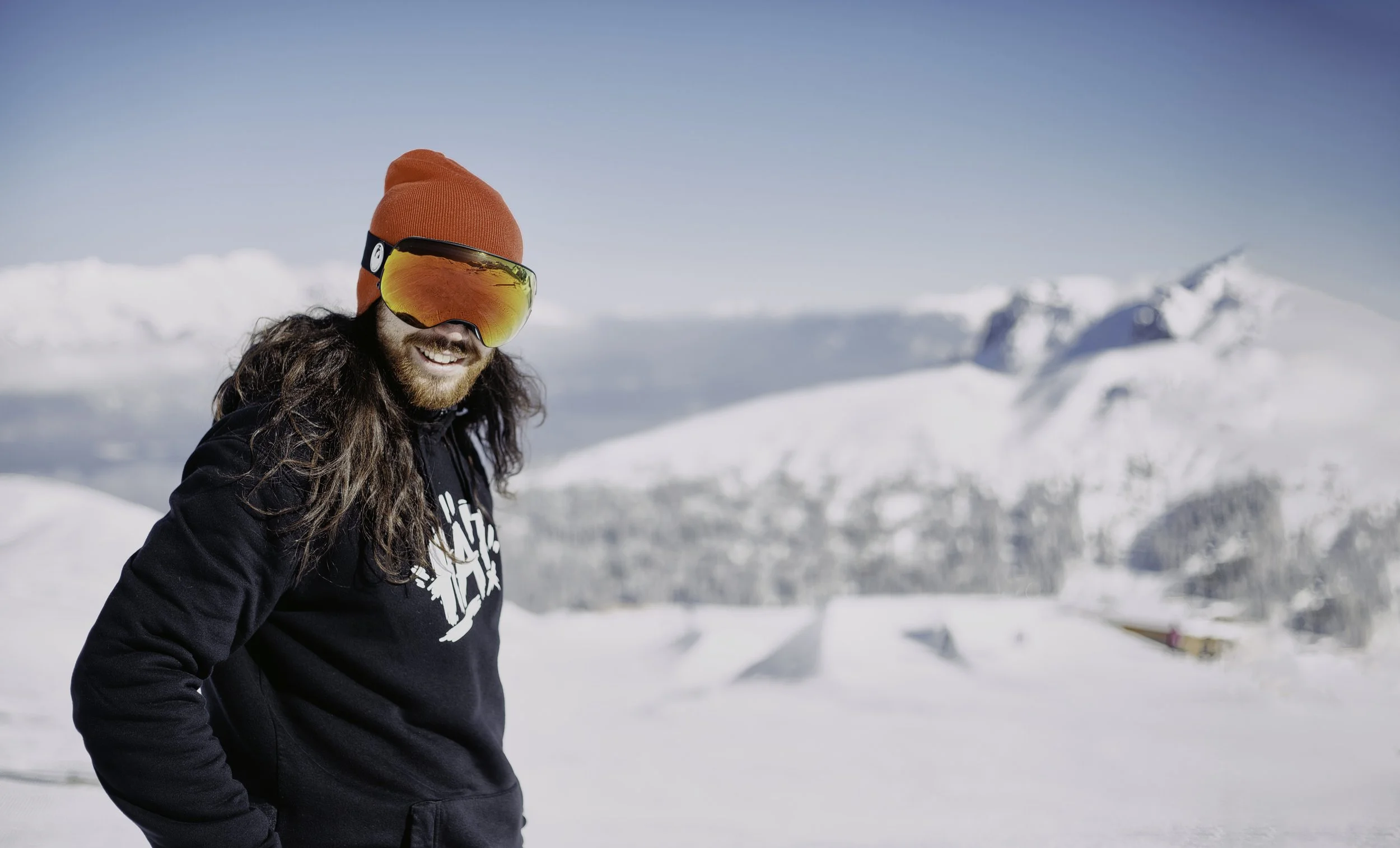 Ein Mann in Skiausrüstung, mit roten Mütze und Skibrille, lächelt in einer verschneiten Berglandschaft.