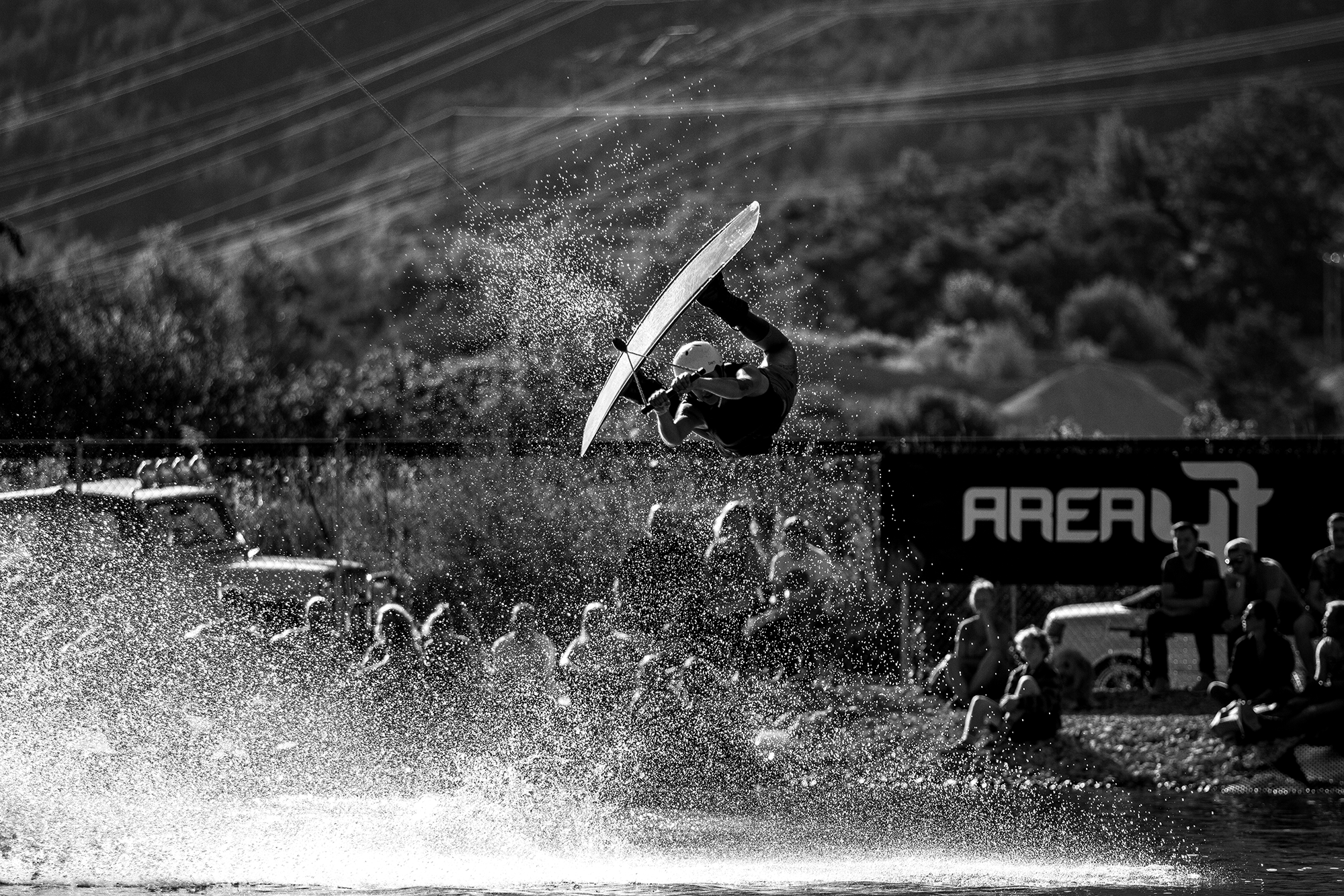 Ein Extremsportler macht einen Salto auf einem Wasser-Skateboard während eines Wettbewerbs, im Hintergrund sind Zuschauer und eine Werbung mit dem Text 'AREALUE' zu sehen.