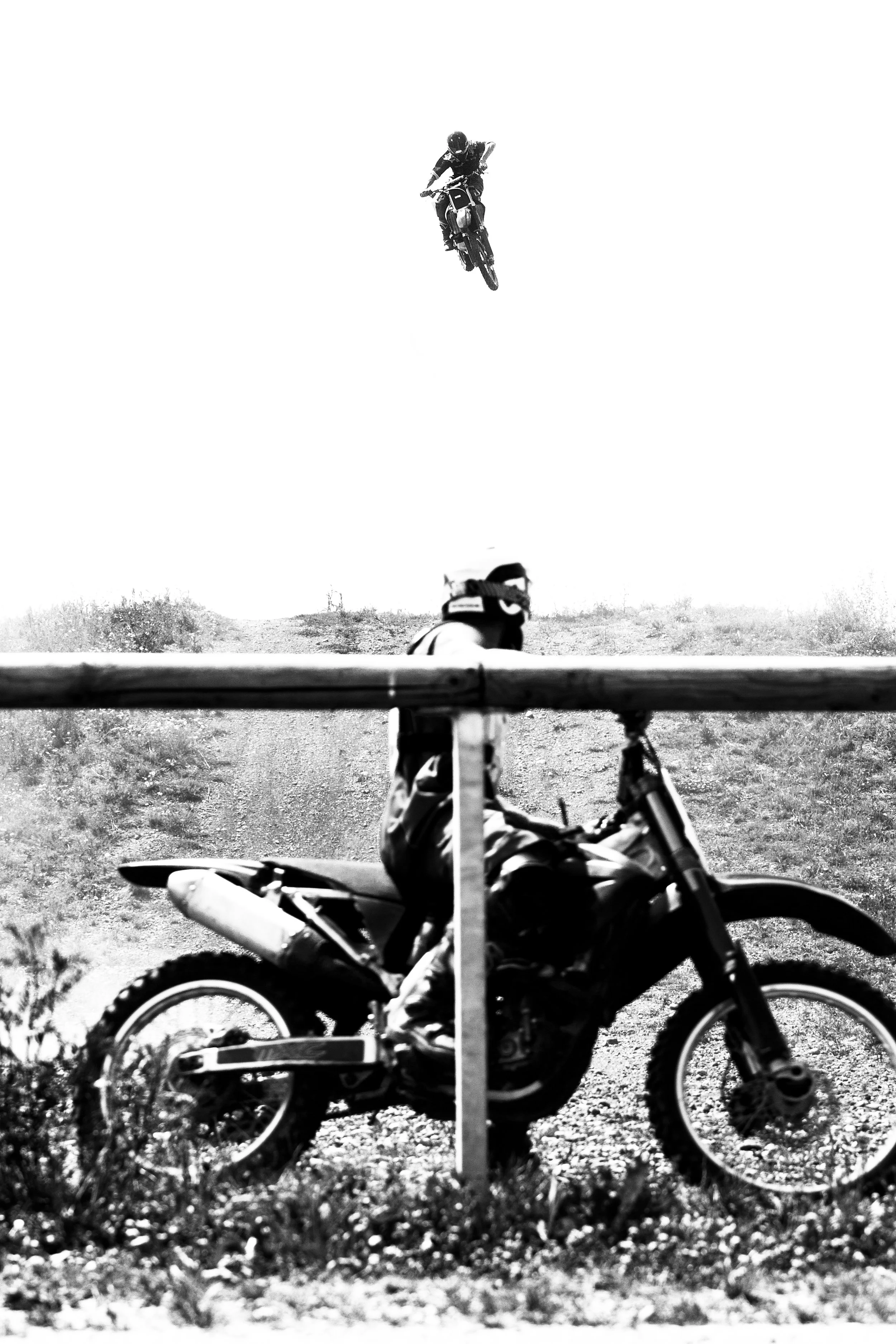 Motorradfahrer sitzt auf einem Motorrad, während ein weiterer Motorradfahrer in der Luft einen Sprung macht, alles in Schwarz-Weiß.