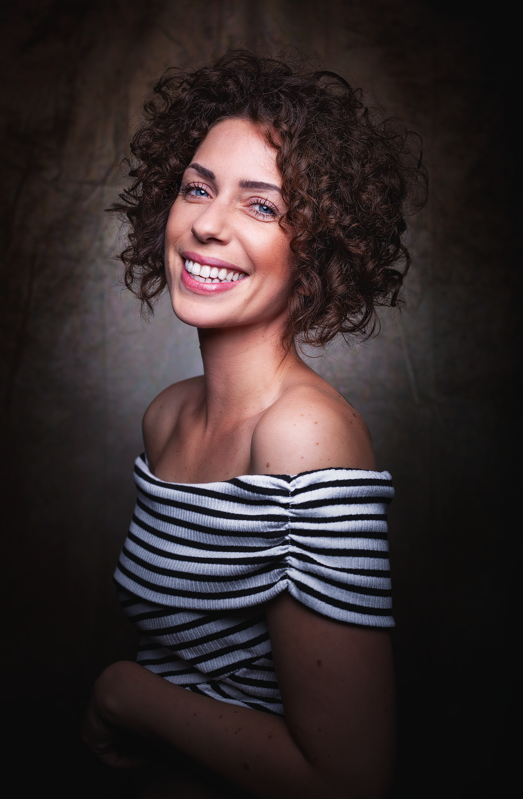 Lächelnde Frau mit lockigen braunen Haaren, in einem schwarzen und weißen gestreiften Oberteil, vor dunklem Hintergrund.
