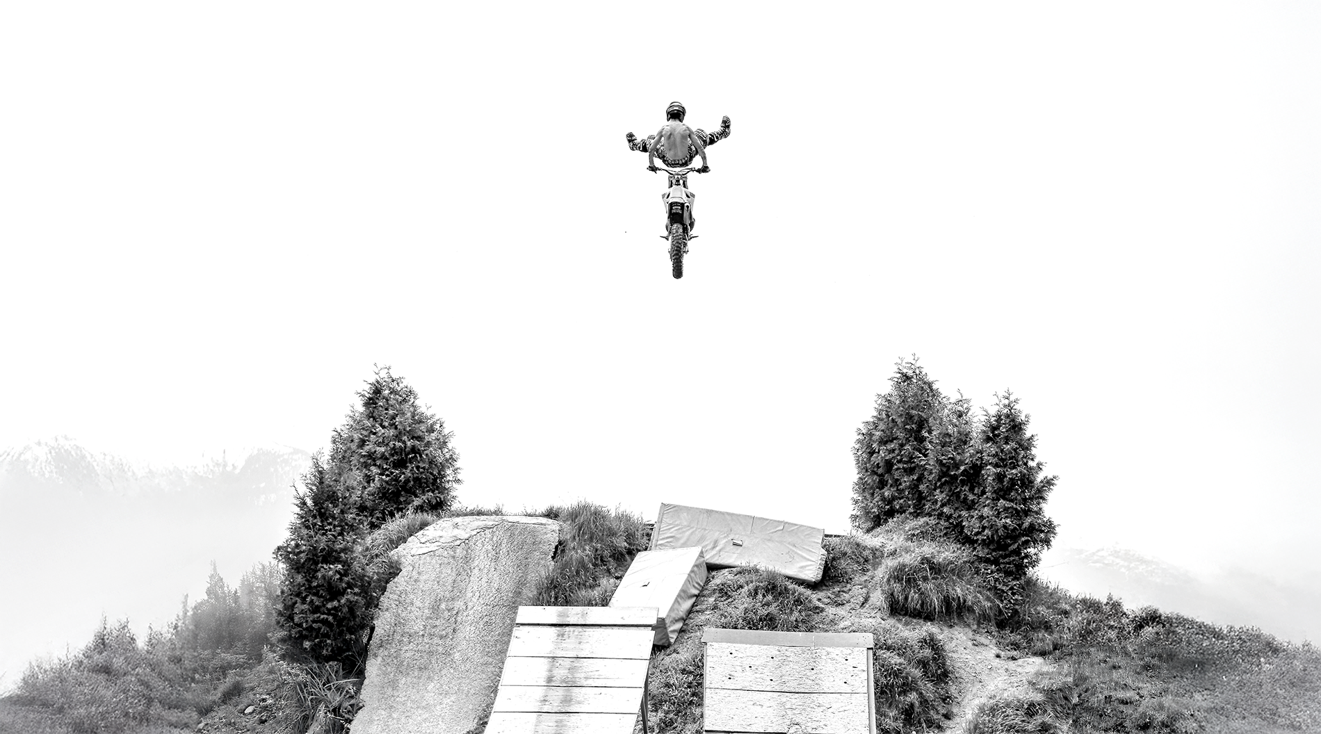 Ein Radfahrer springt mit seinem Mountainbike von einer Rampe auf einen Hügel in der Natur, im Hintergrund sind Bäume und Berge zu sehen.