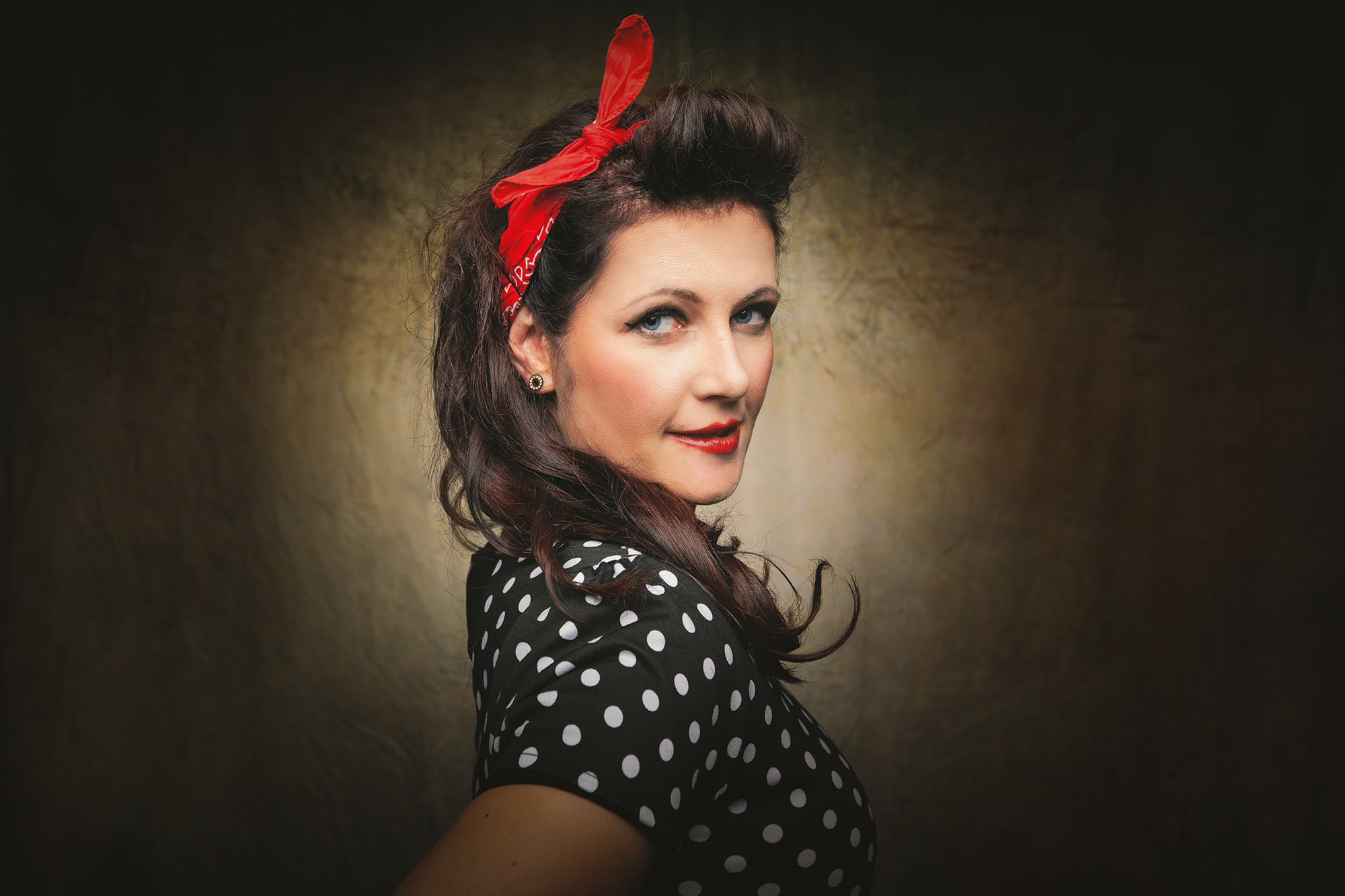 Frau im Retro-Style mit schwarzen Kleid mit weißen Punkten, roter Haarband, roten Lippen, dunklem Haar, im Porträt mit dunklem Hintergrund.