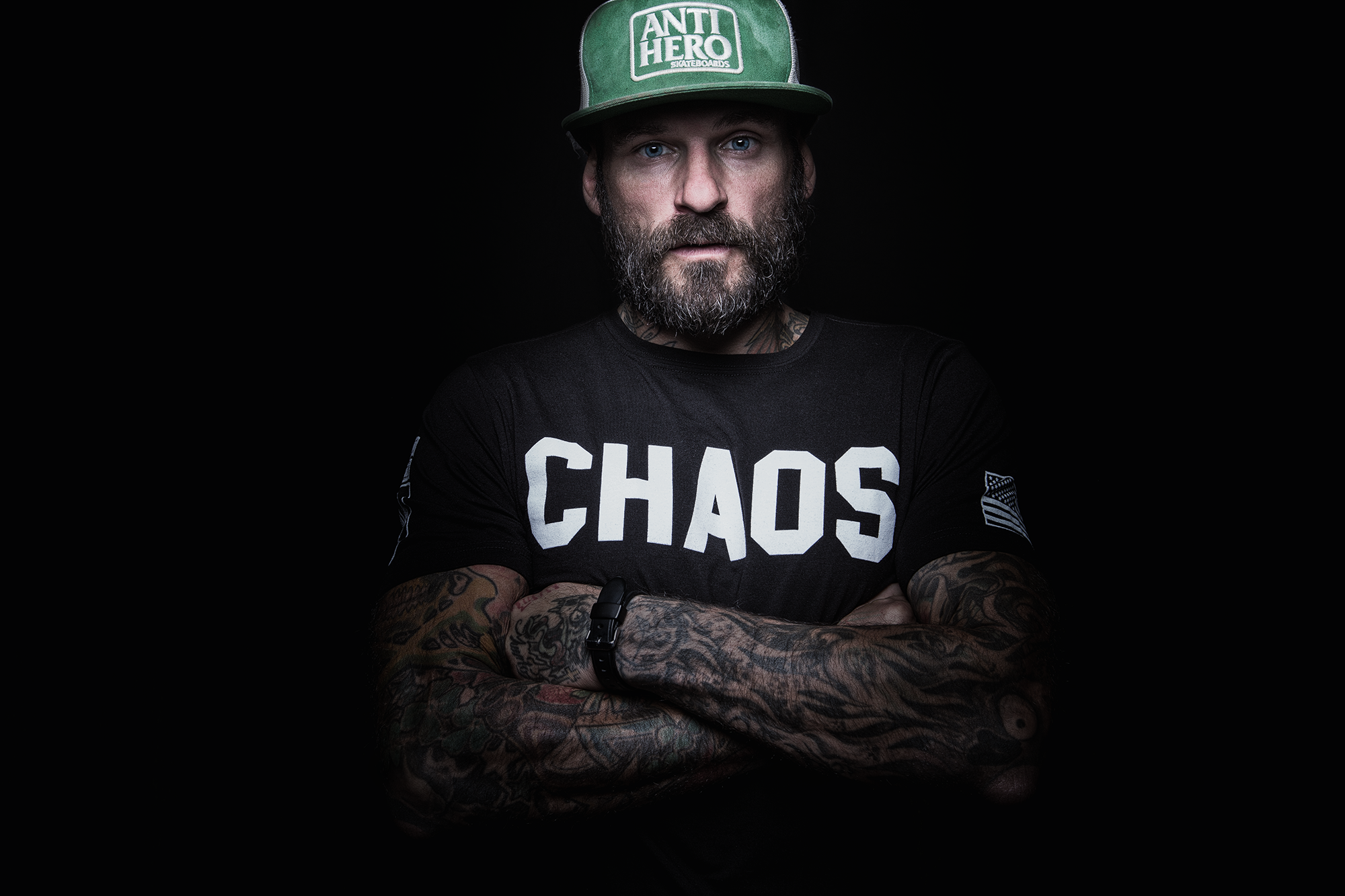 Junger Mann mit Tattoos, blauer Augen, grauem Bart und schwarzen T-Shirt mit weißen Buchstaben, trägt eine grüne Cap mit Aufdruck 'ANTI HERO' und 'SKATEBOARDS', steht vor dunklem Hintergrund.