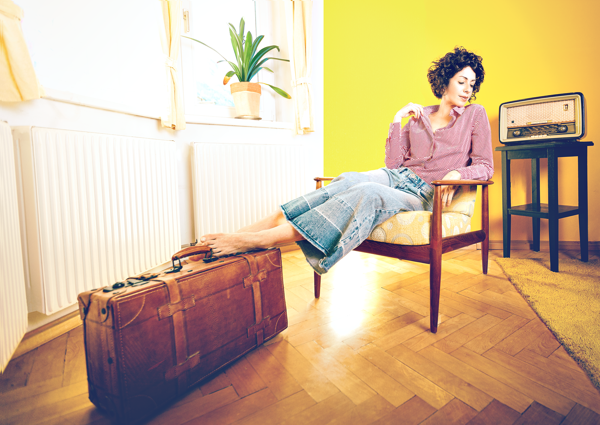 Eine Frau sitzt entspannt in einem gemütlichen Wohnzimmer auf einem Holzstuhl mit gepolstertem Sitz. Ihre Beine sind auf einem alten, braunen Koffer mit Rollen und Griff gelegt. Neben ihr steht ein schwarzer Beistelltisch mit einem vintage Radio. Das