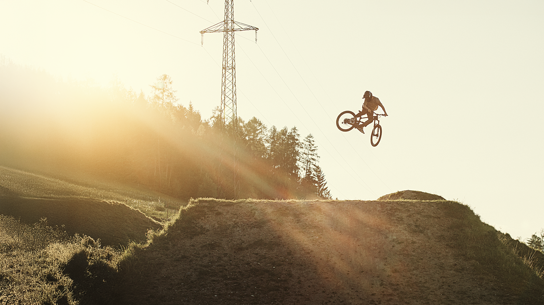 Mountainbiker springt bei Sonnenuntergang auf einem Dirt-Hügel in der Natur. Im Hintergrund sind Bäume und ein Strommast sichtbar.