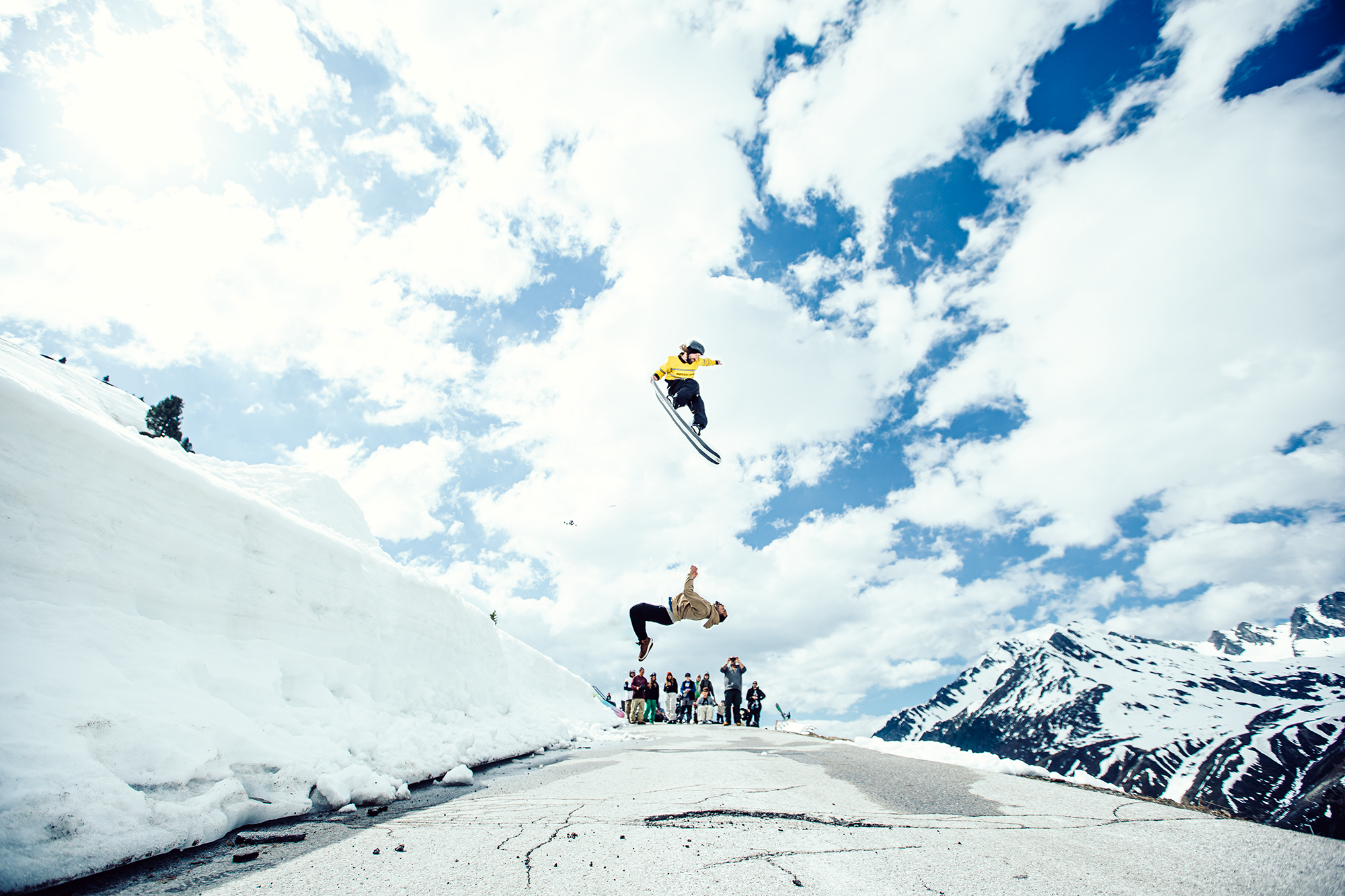 Ein Skifahrer springt in der Luft auf einer schneebedeckten Straße mit Bergen im Hintergrund, während eine Gruppe von Menschen zuschaut.
