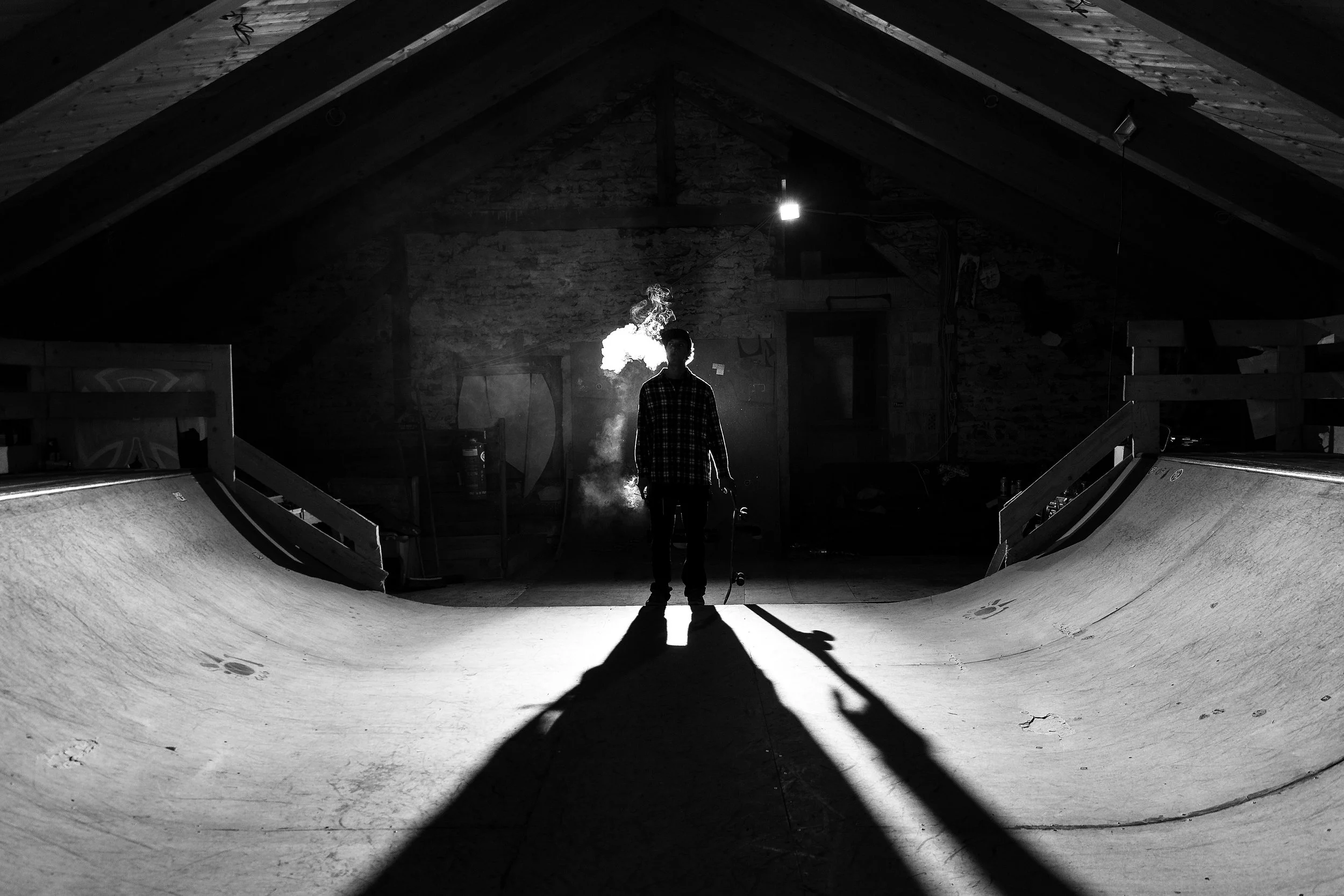 Junge steht im Skatepark mit rauchender Zigarette, Schatten auf dem Boden, dunkler Raum mit Holzdecke, im Hintergrund.