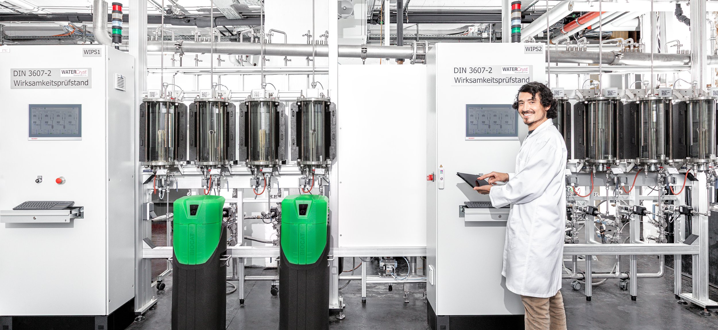 Mann in weißem Laborkittel, der mit einem Tablet vor chemischer Labor- oder Wasseraufbereitungsanlage steht, in einem technischen Raum.