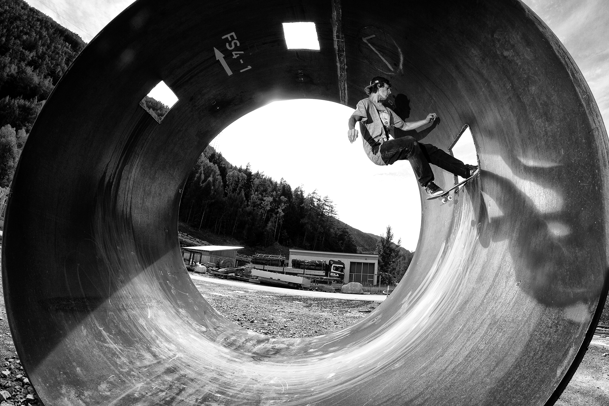 Ein Skater fährt durch ein großes Metallrohr im Freien, umgeben von Bäumen und Hügeln.