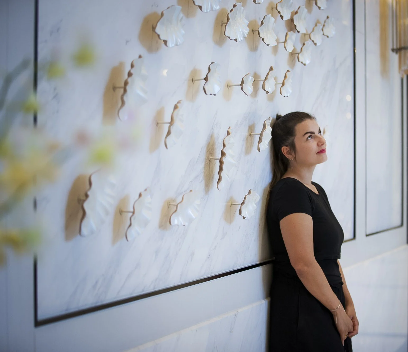 Eine Frau lehnt an einer Wand mit weißen, kunstvollen Skulpturen. Sie trägt ein schwarzes Kleid und schaut nach oben.