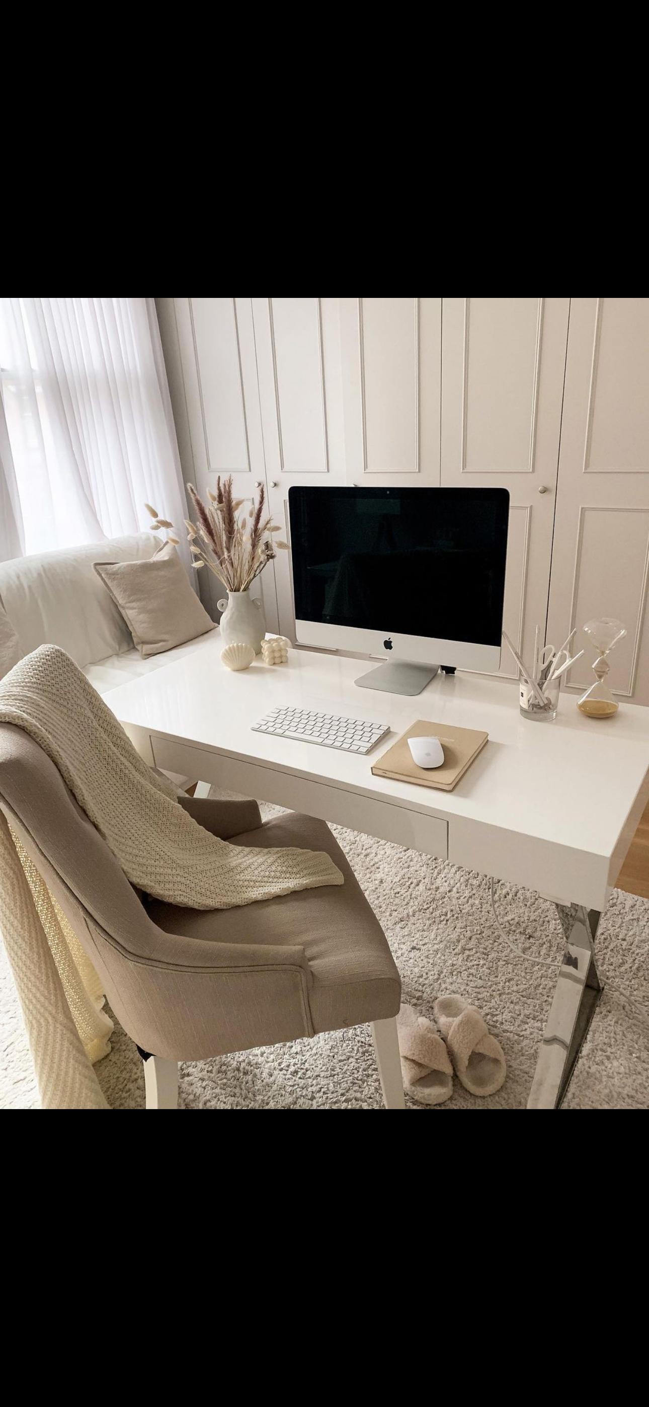 A modern, minimalistic home office with a white desk, beige chair with a knitted throw, desktop computer, keyboard, mouse, notebooks, and decorative items like a vase with dried flowers, an hourglass, and small sculptures. The room has white panel cabinets and sheer curtains.