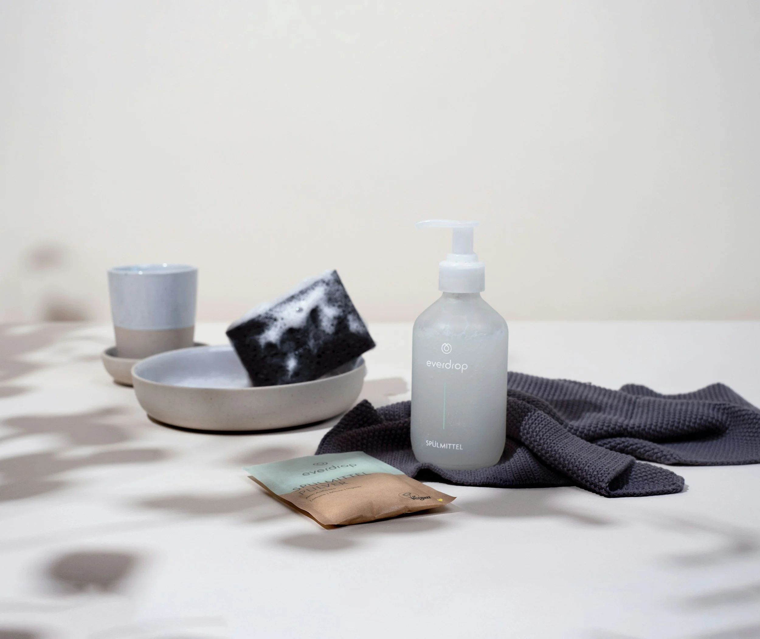 A bottle of hand soap labeled 'everdrop' with a pump dispenser is on a dark gray cloth on a white surface. Next to it is a small packet with the same brand and the word 'PULVERIZER.' In the background, there is a white dish with a black sponge covered in soap suds, and another small white cup with a matching saucer.