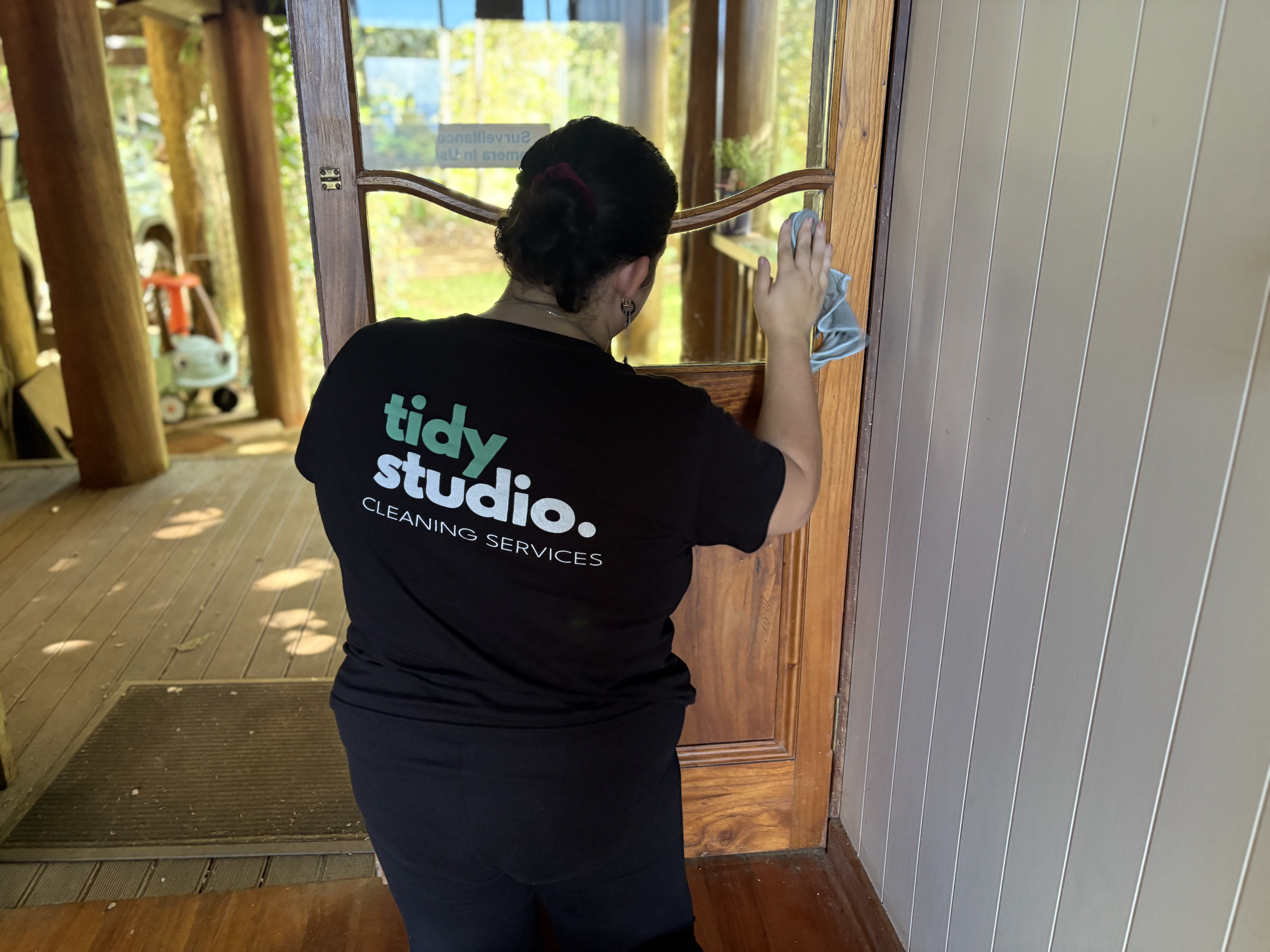 A woman wearing a black shirt with the words 'tidy studio' is cleaning a wooden window frame with a cloth in a cozy interior space with wood flooring and vertical-paneled walls. Outside, there is greenery and a small toy car.