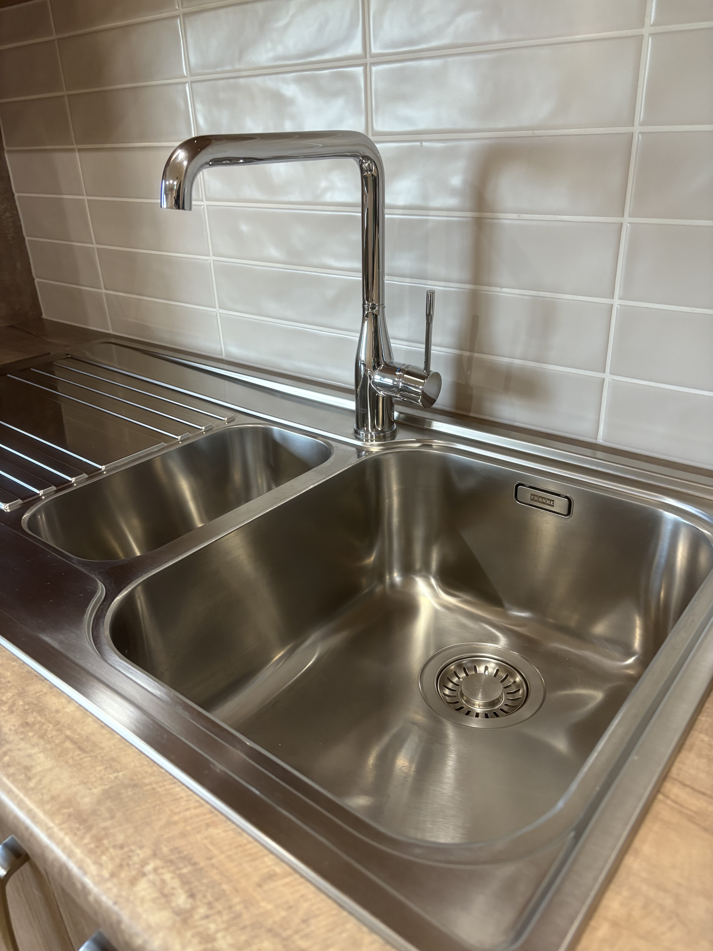 Stainless steel kitchen sink with two basins, a modern chrome faucet, and a wooden countertop, with beige tiled backsplash.