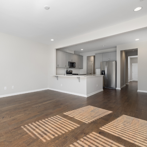 Empty living room with hardwood floors and kitchen in the background, sunlight casting shadows through window blinds.